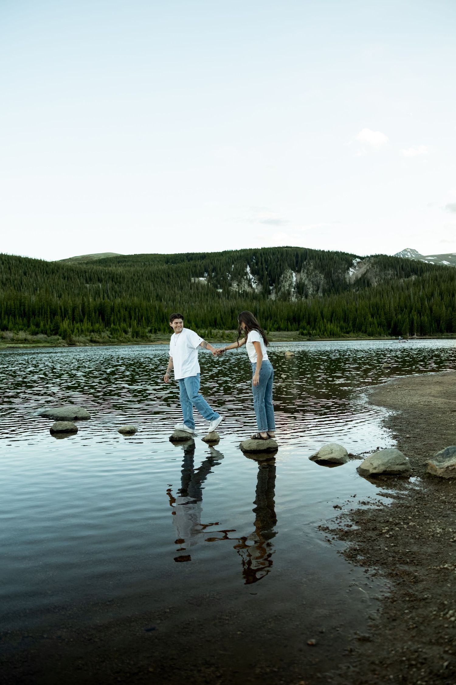 Couple holding hands while stepping on rocks in a calm mountain lake, with forested hills reflected in the water.