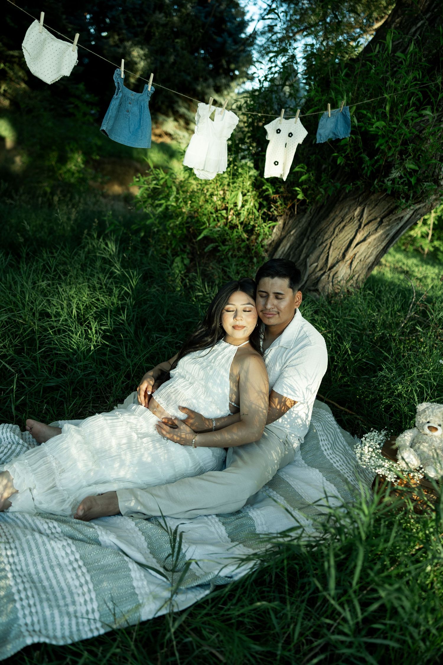 Couple sitting on blanket in lush green park, pregnant woman in white dress, baby clothes hanging on line above them.