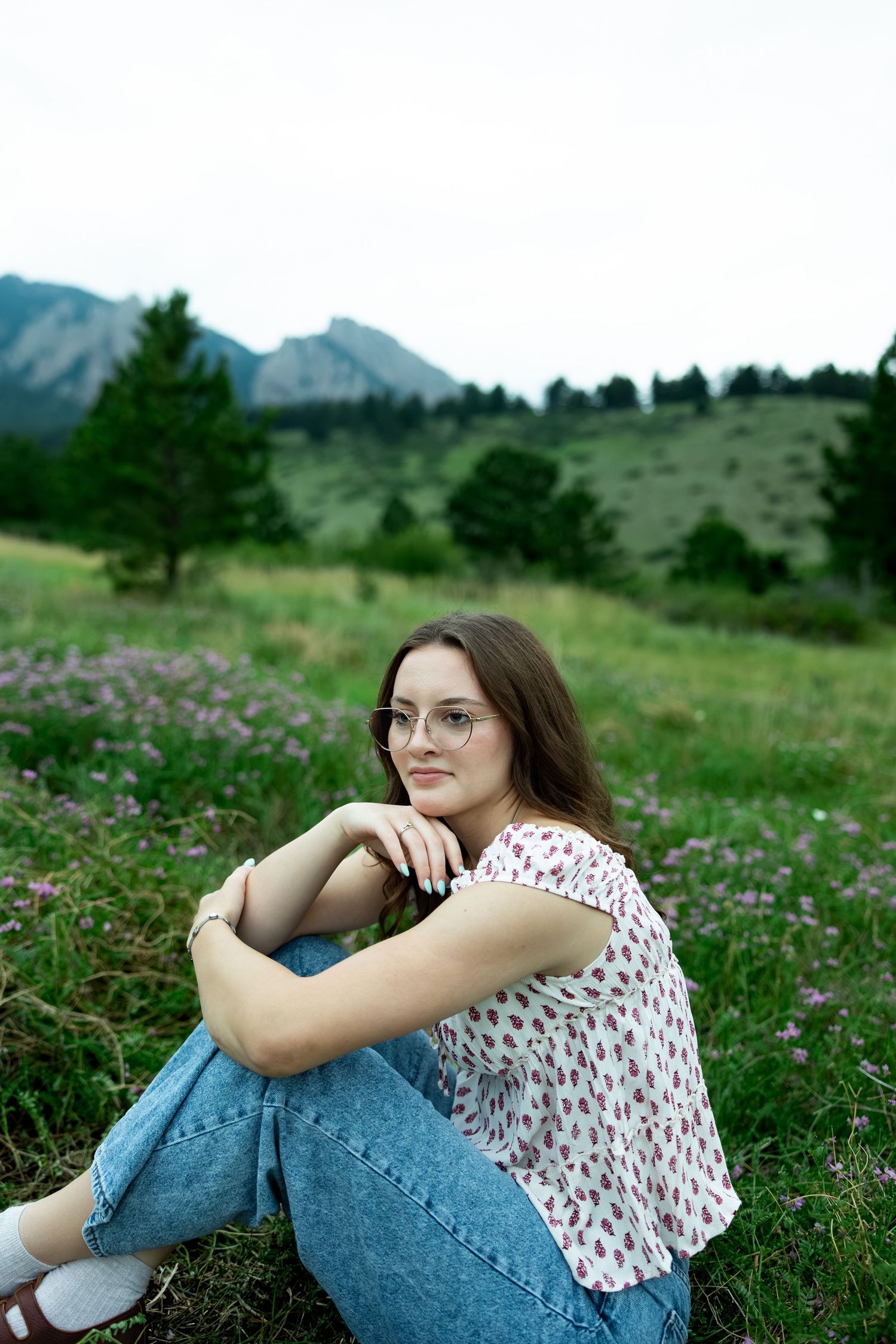 Young woman in floral top and jeans sitting in a mountain meadow with wildflowers and pine trees.