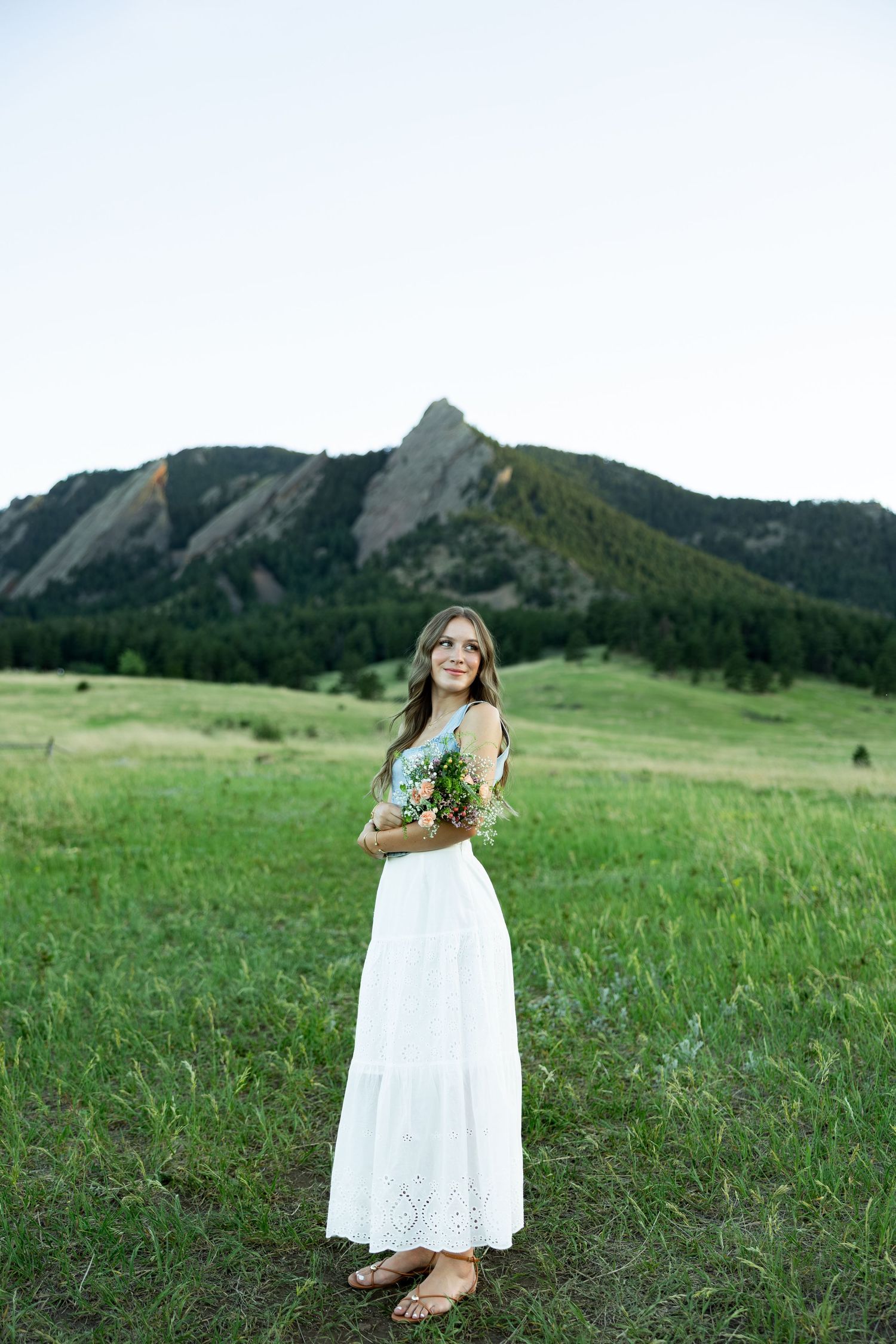 Young woman in white maxi dress holding wildflowers in a green meadow with the Flatirons mountains in Boulder, Colorado.