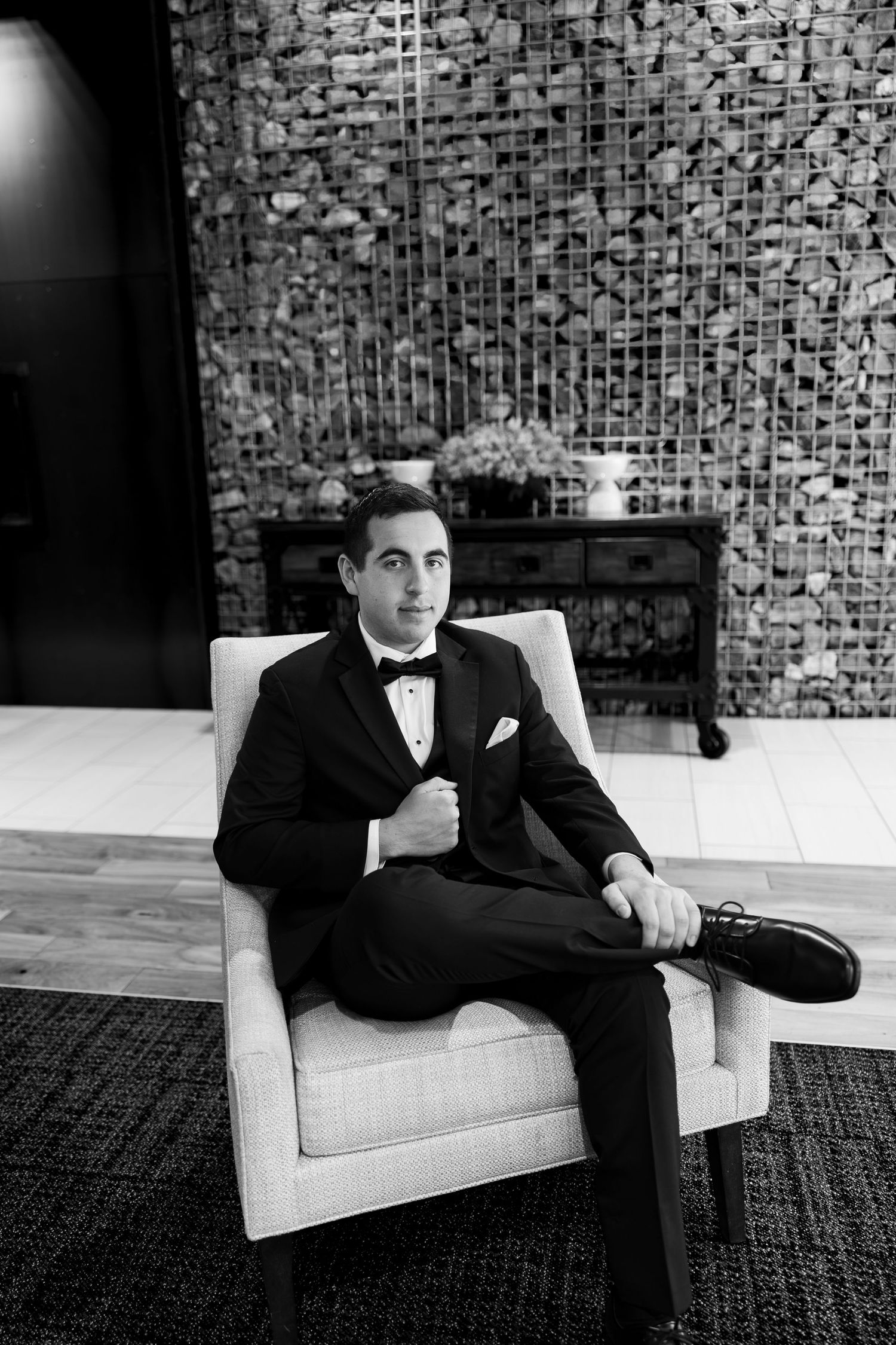 A dapper groom in a black tuxedo and bow tie sits confidently in a white chair against a stone wall backdrop.