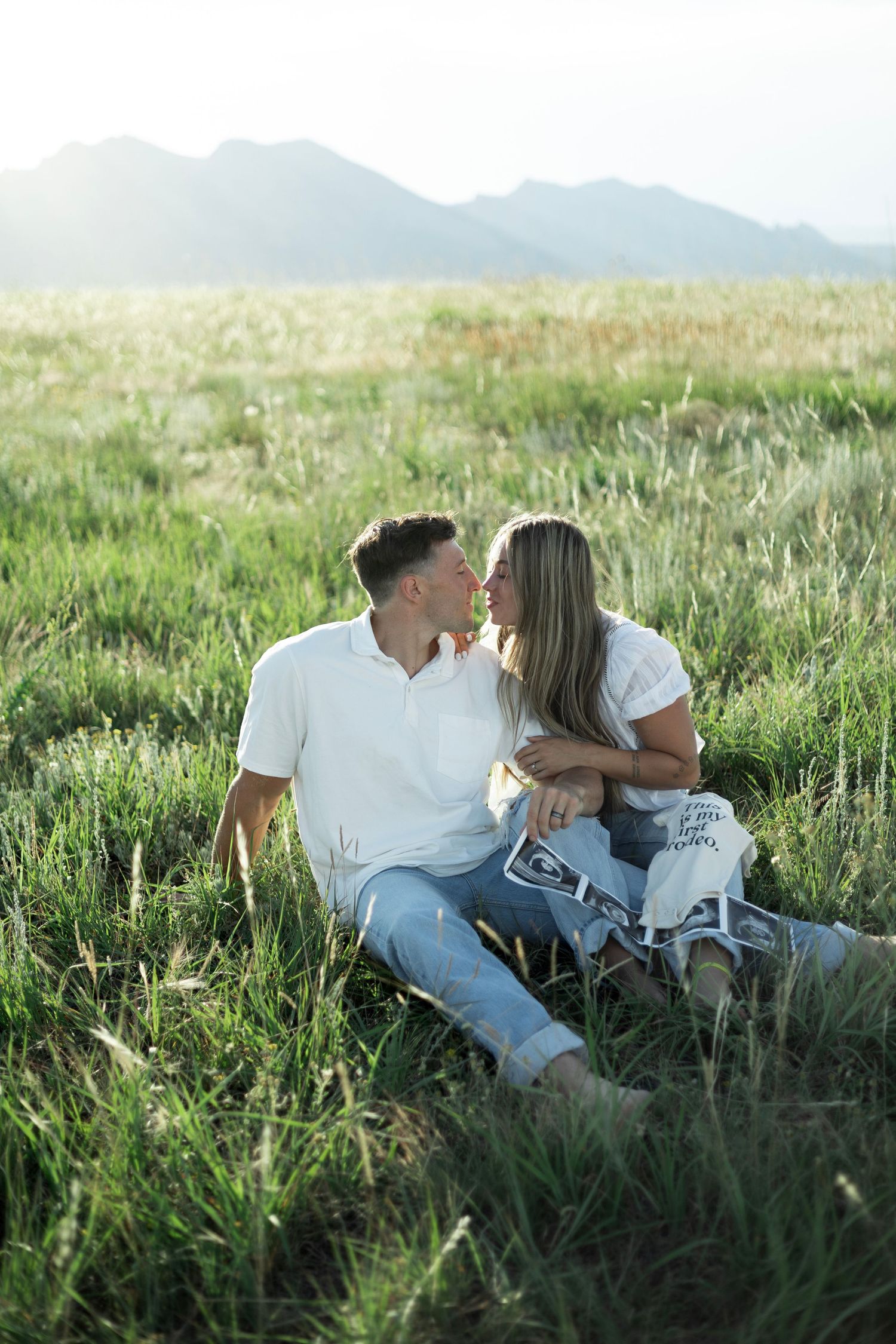A couple sits together intimately in a grassy field with mountains visible in the misty background.