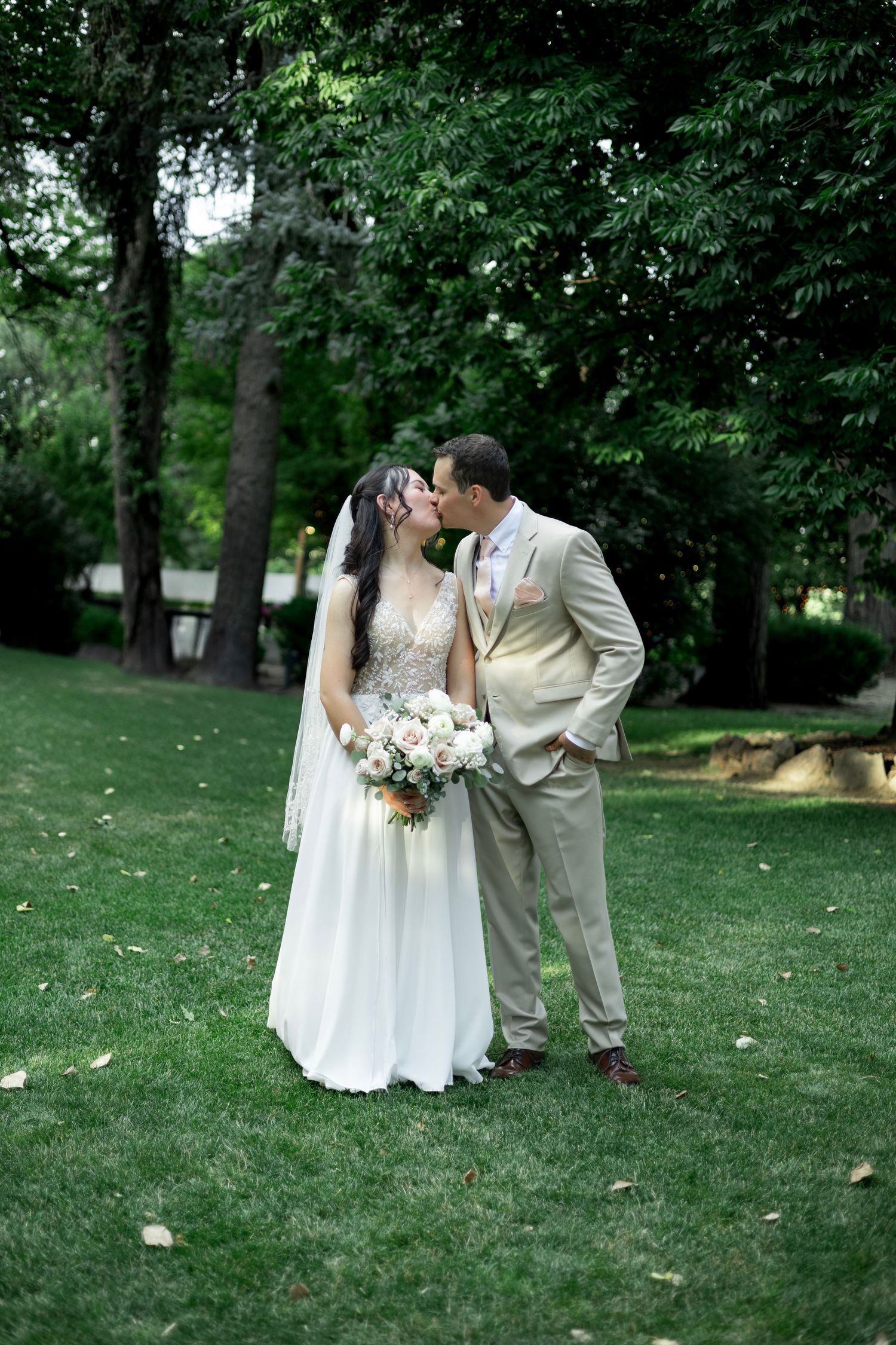 A couple embraces on their wedding day in a lush green garden setting, with the bride holding a white floral bouquet.