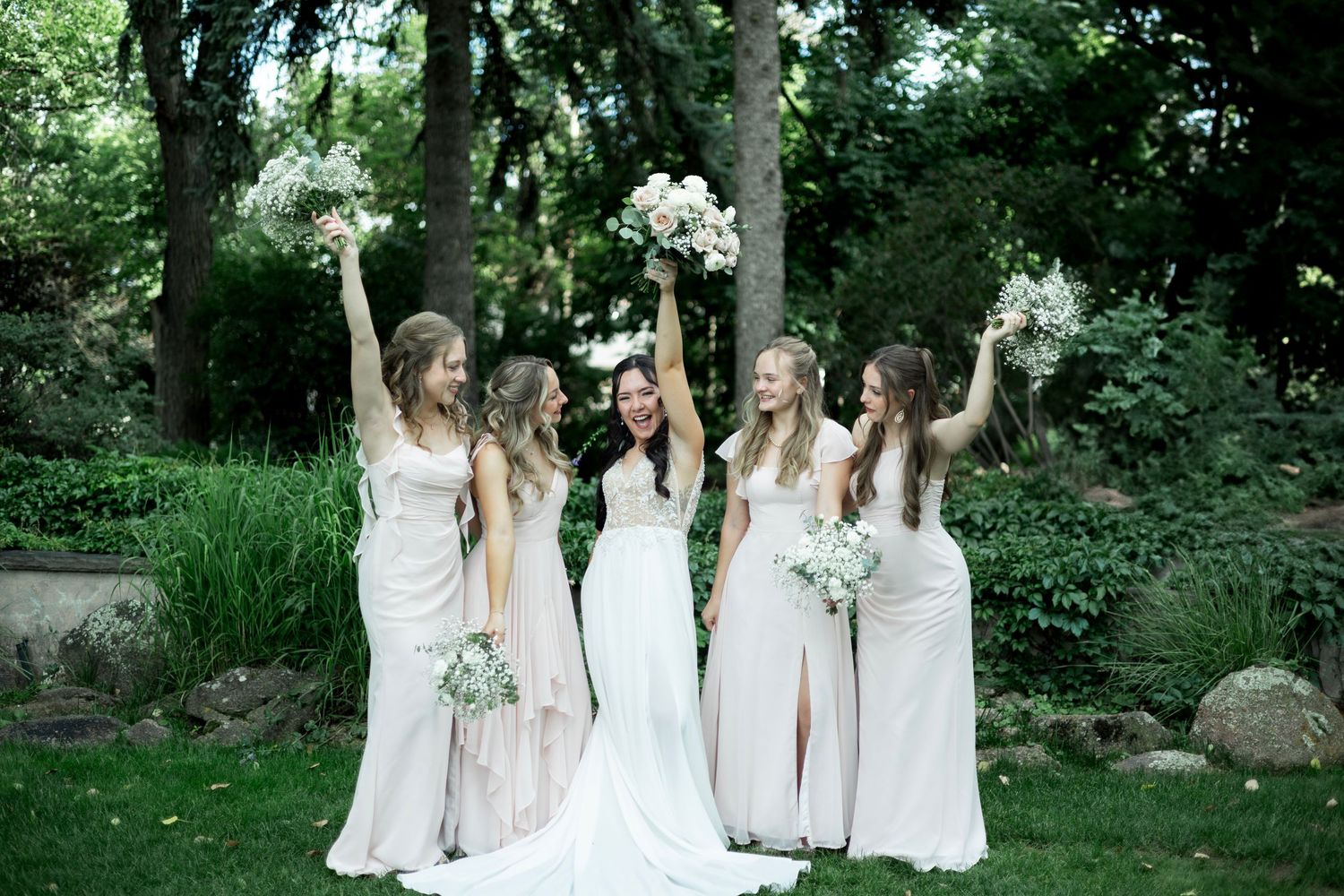A wedding party wearing white dresses holds bouquets aloft in celebration while standing in a lush garden setting.