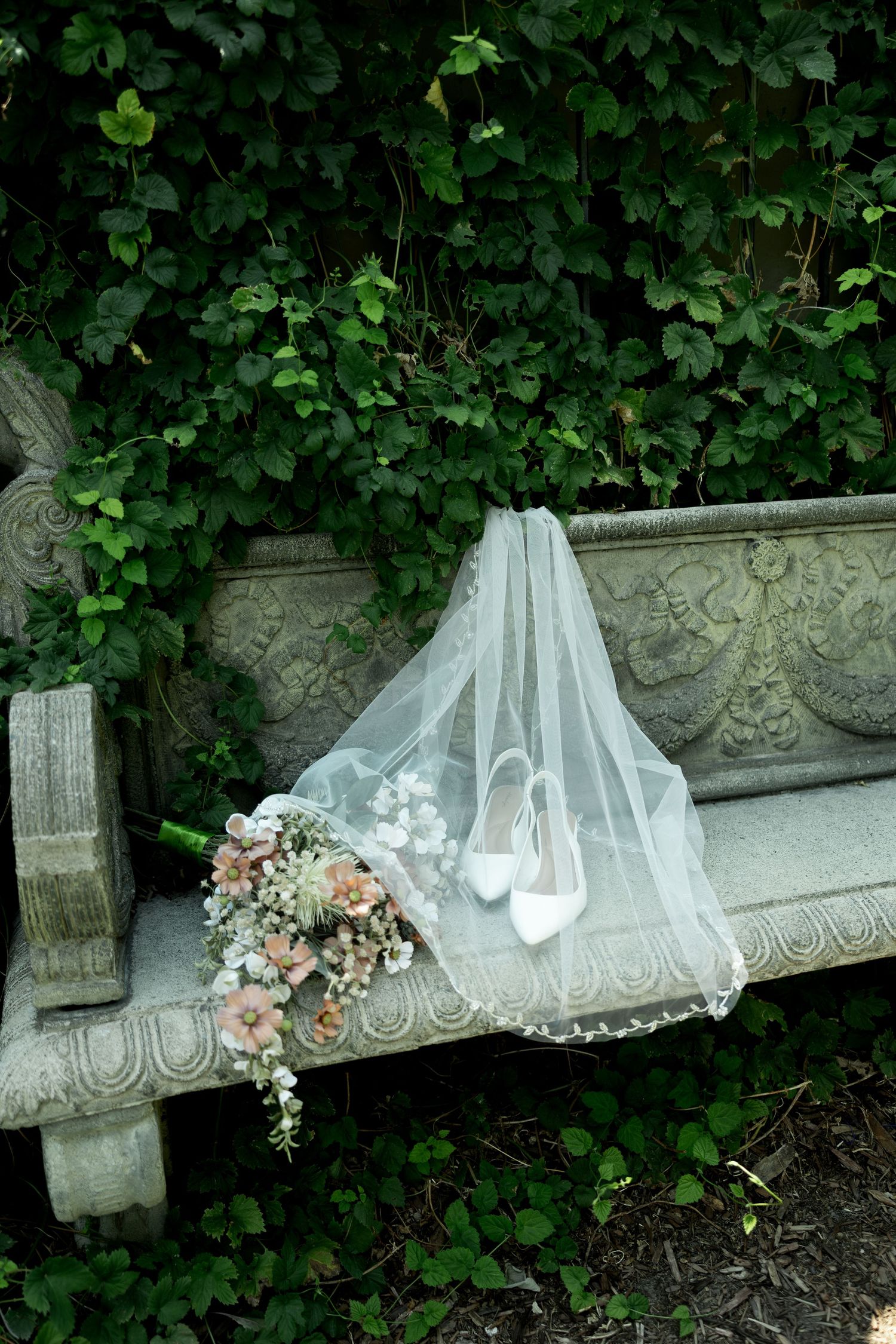 Bridal veil, white heels, and pink floral bouquet arranged on a stone garden bench surrounded by lush ivy.
