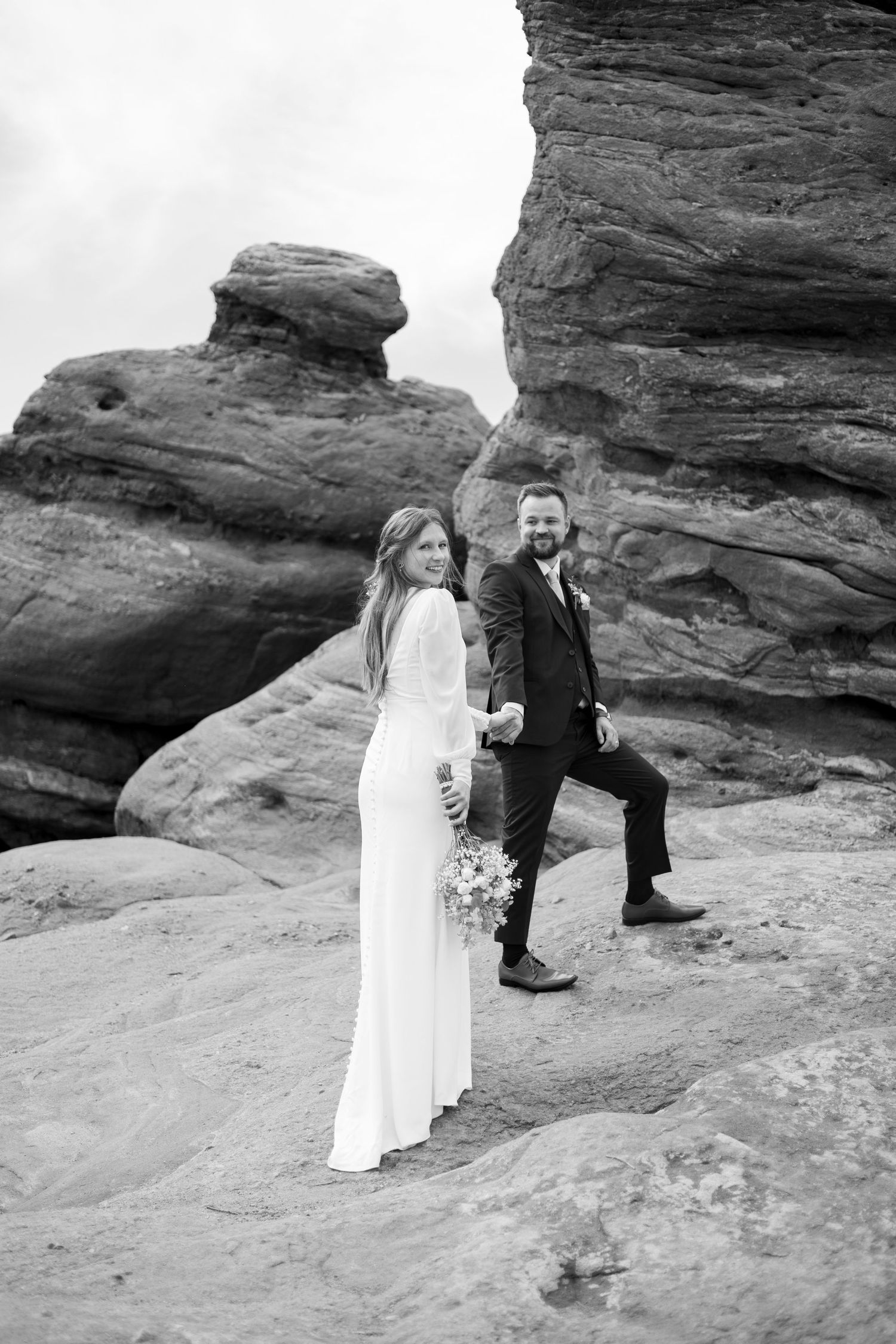 A couple in wedding attire embraces while standing on red rock formations in a desert landscape.