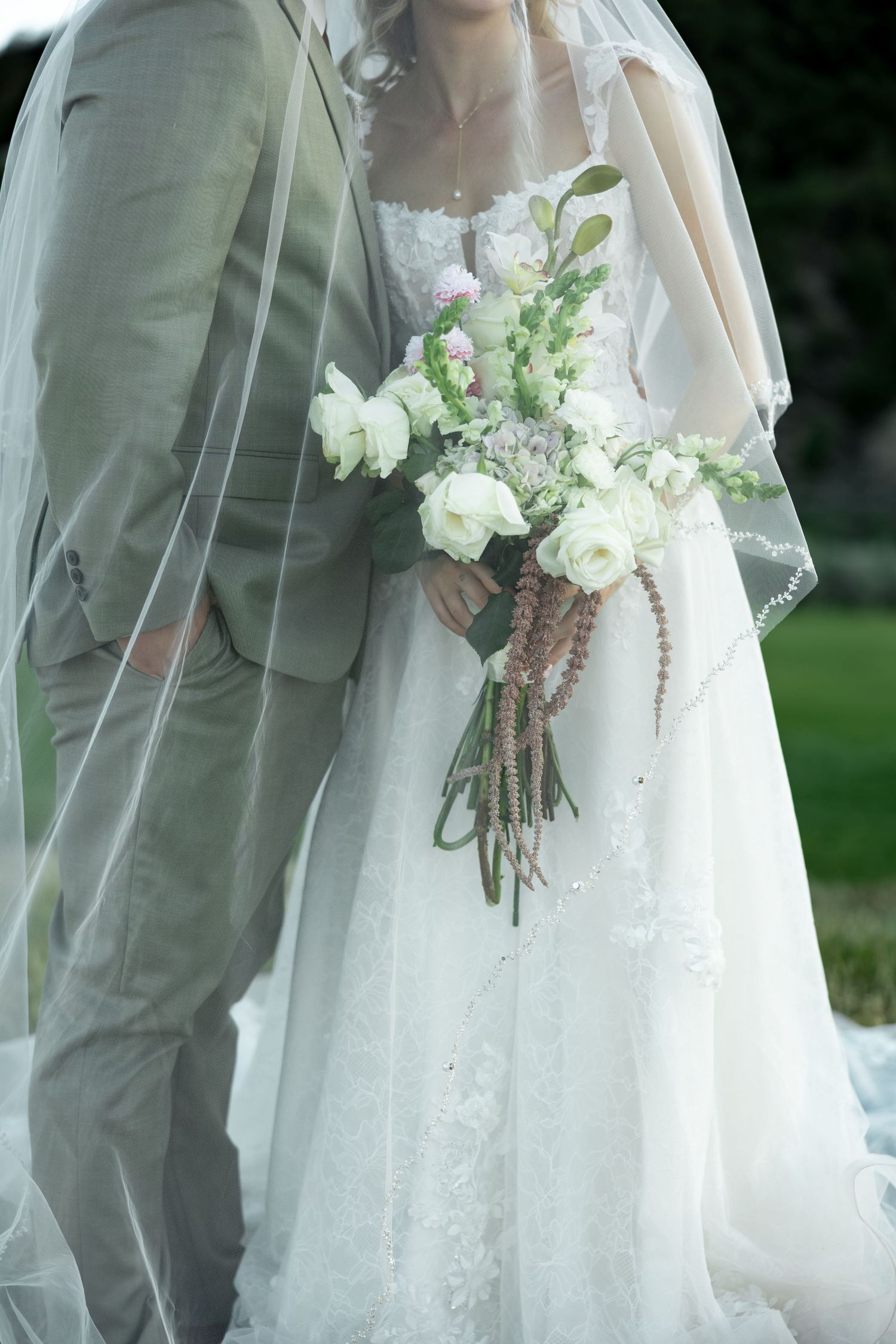 Bride in lace gown holding white floral bouquet stands beside groom in sage suit, veil flowing softly outdoors.