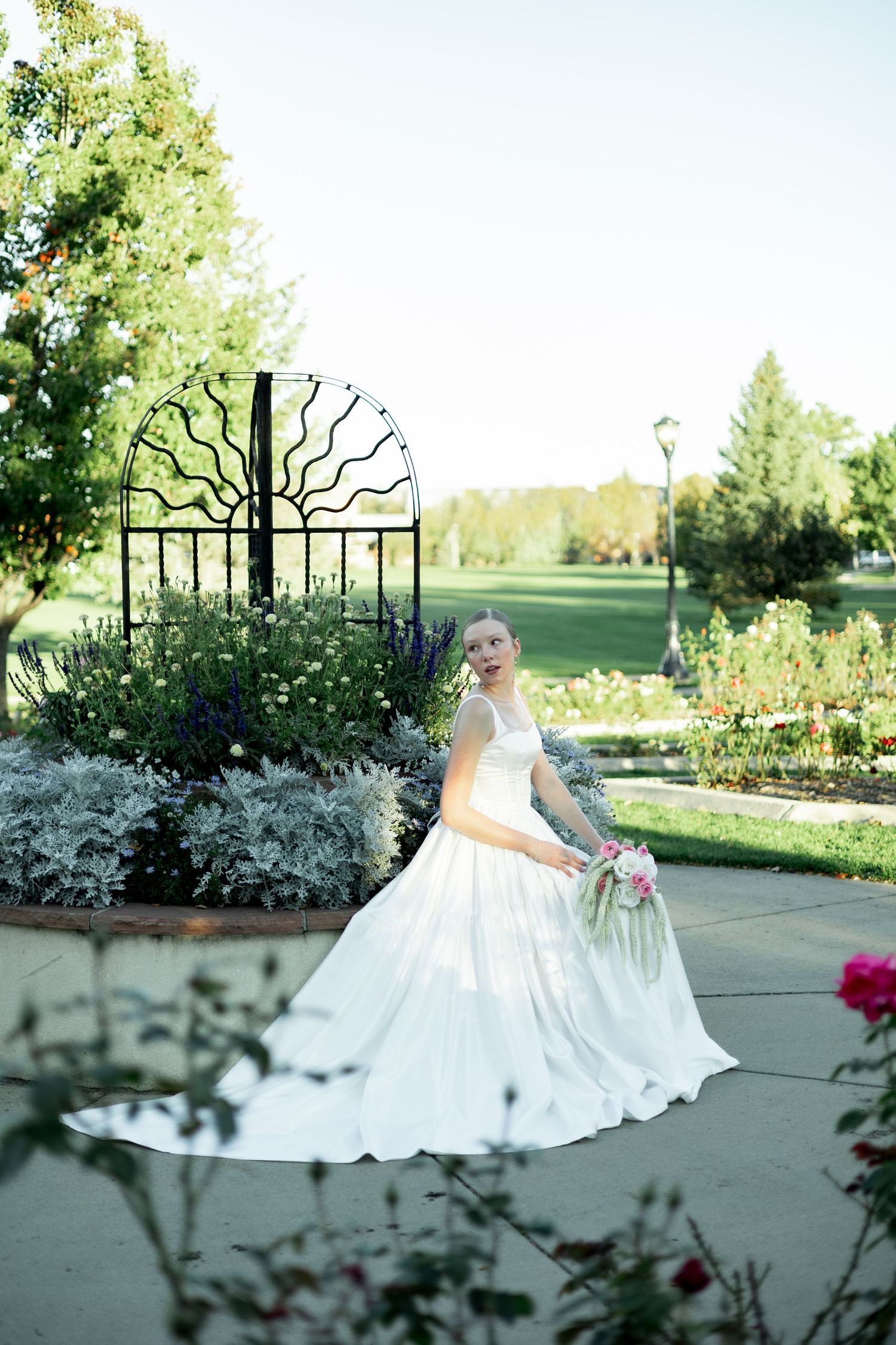 A white bridal gown billows elegantly against a backdrop of blooming garden flowers and greenery.