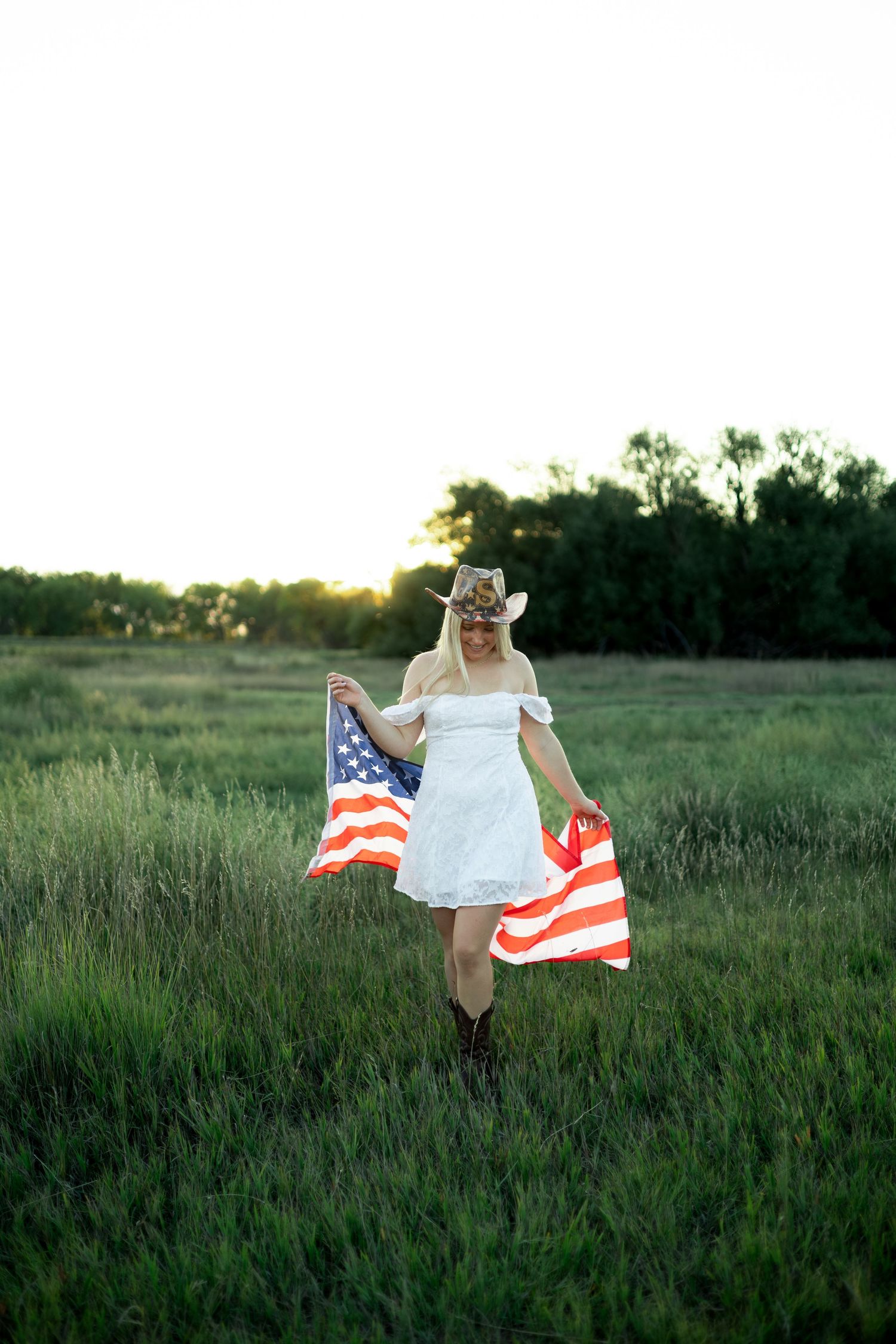 A person runs through a grassy field at sunset while holding an American flag in a flowing white dress.