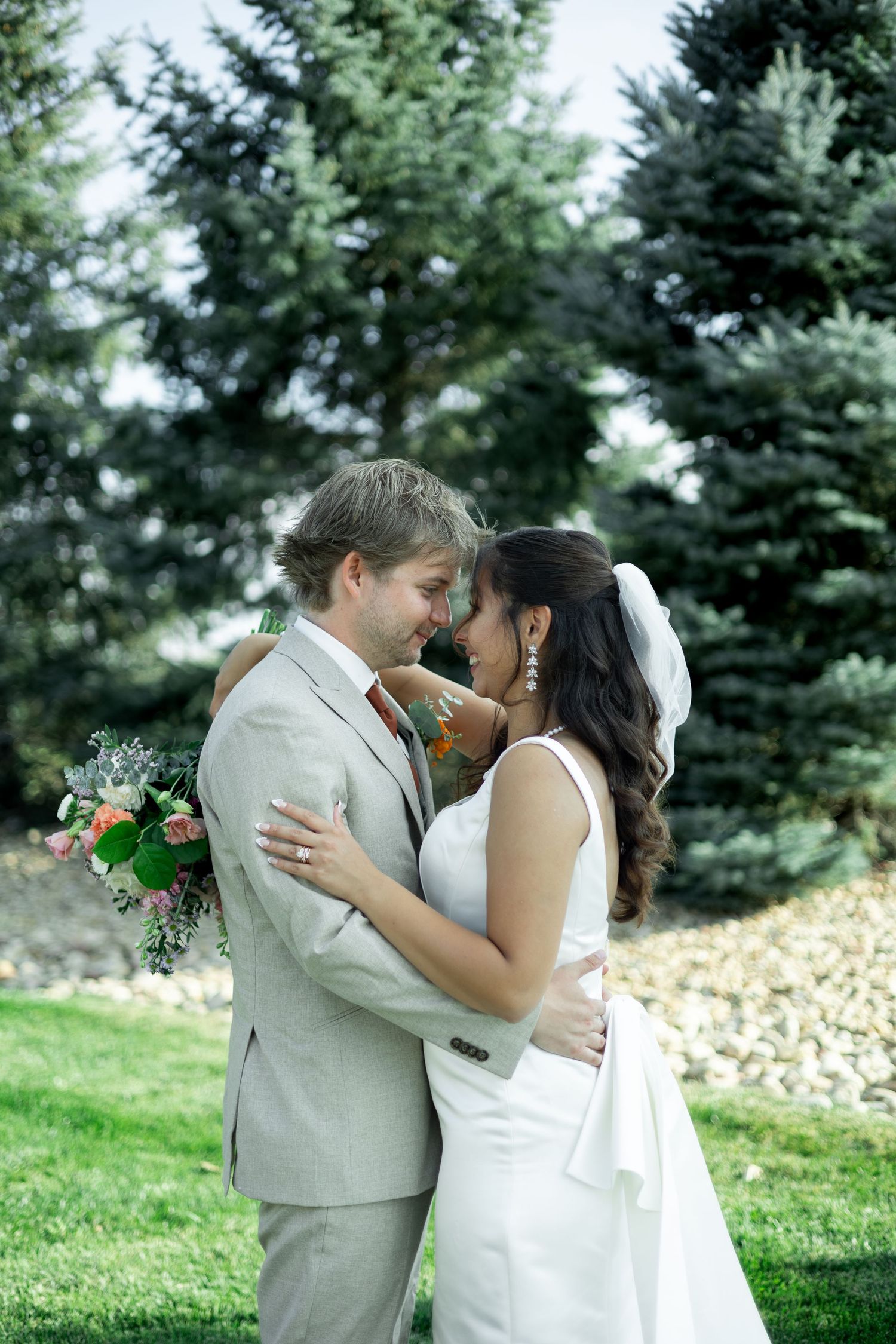 A couple shares a romantic kiss outdoors with evergreen trees in the background during their wedding celebration.