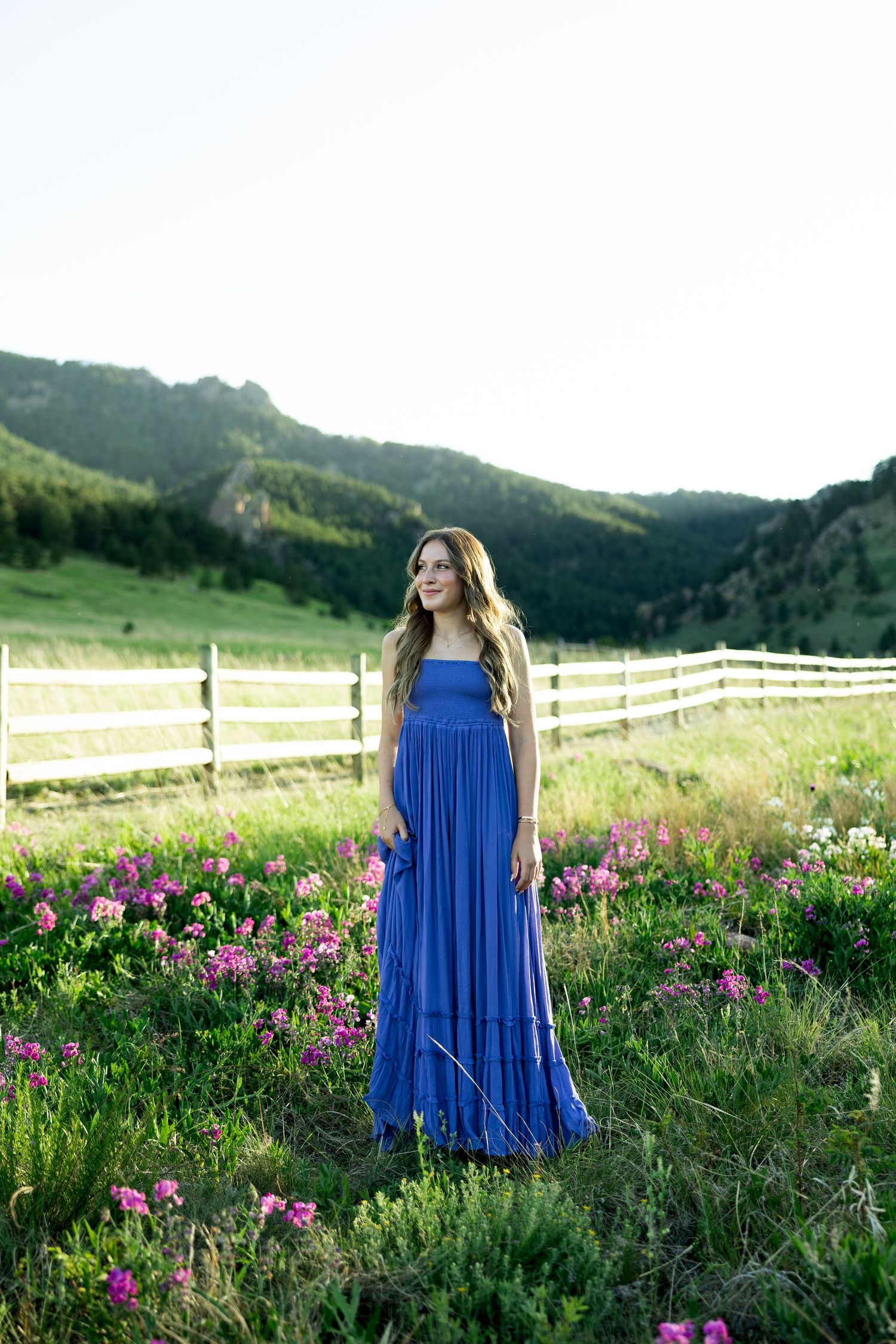Woman in blue maxi dress standing among pink wildflowers with green mountains and white fence.