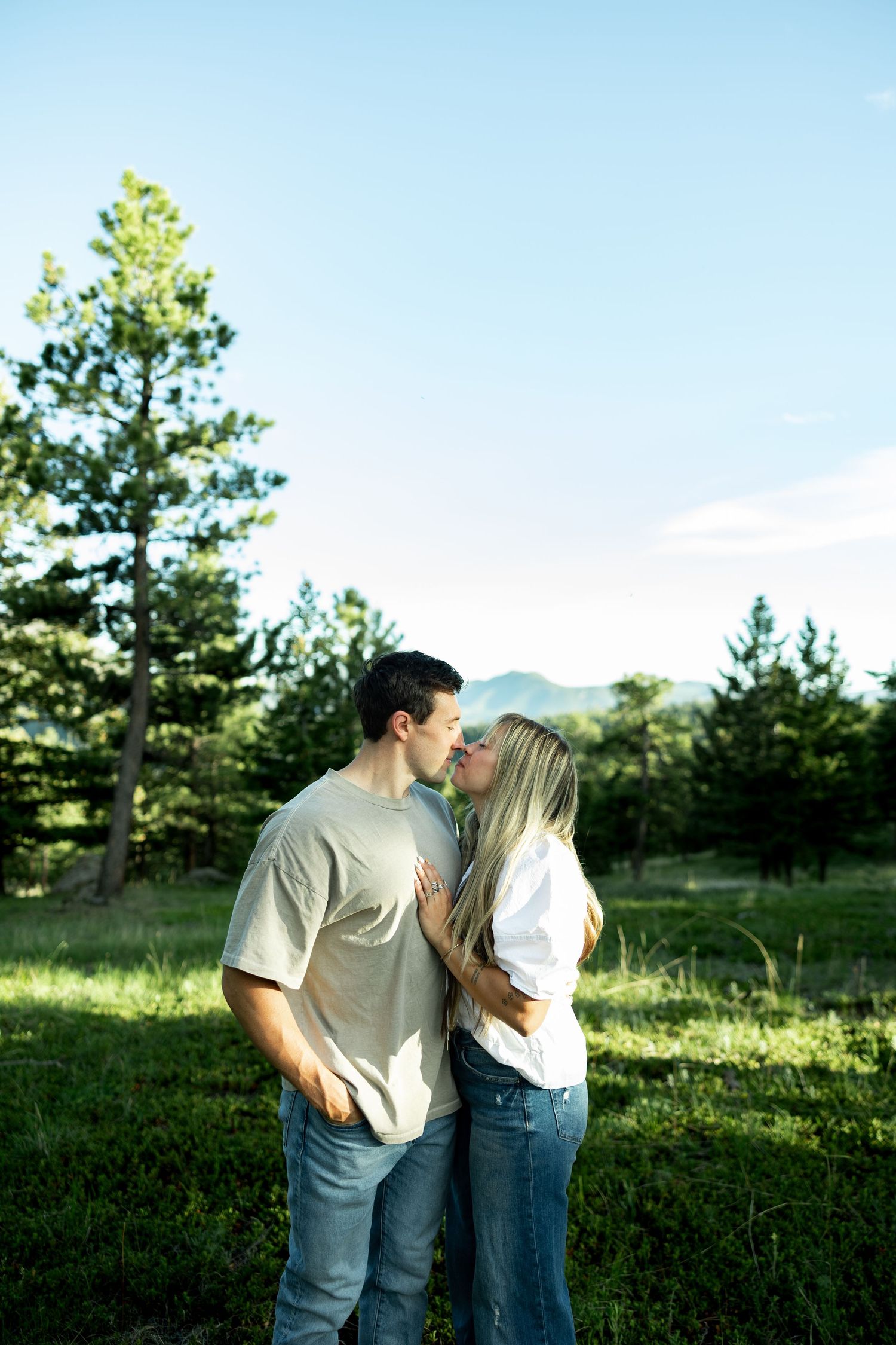 A couple shares a romantic kiss in a lush green meadow surrounded by pine trees and mountains under a clear blue sky.