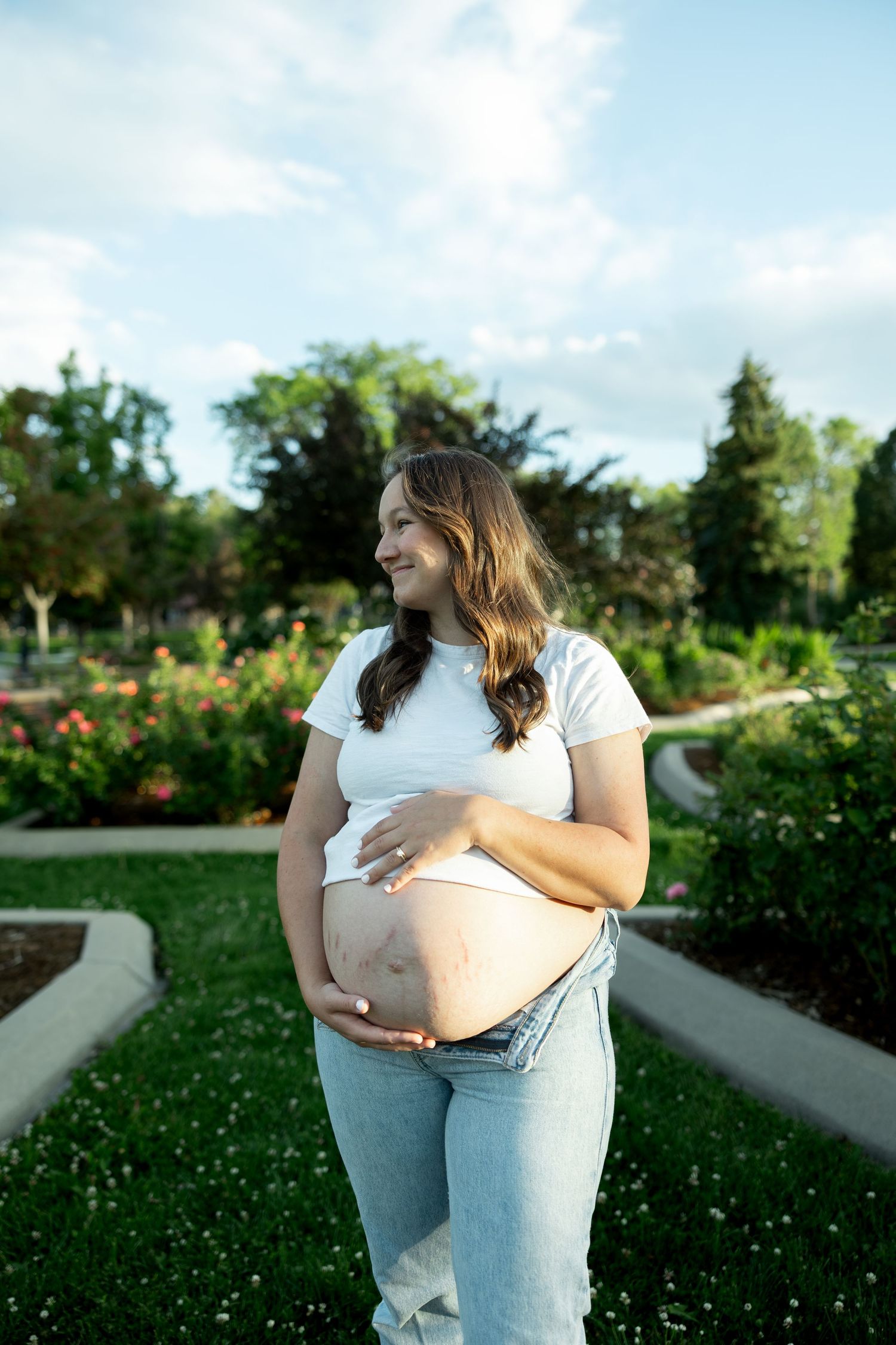 Smiling pregnant woman in white tee and jeans cradling her baby bump outdoors in a lush green park.
