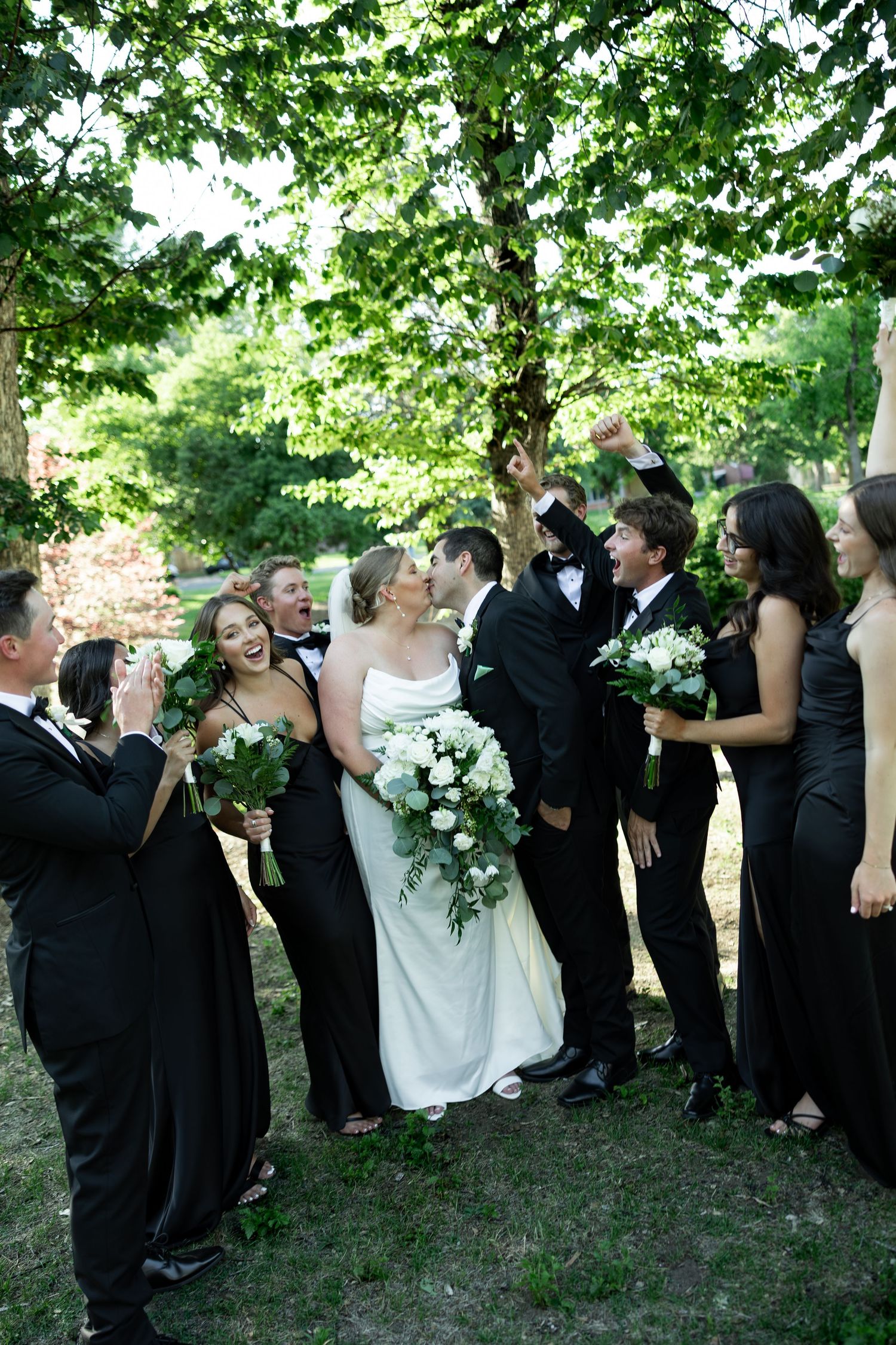 A joyful wedding party in black attire cheers as the bride and groom share a kiss outdoors under lush green trees.