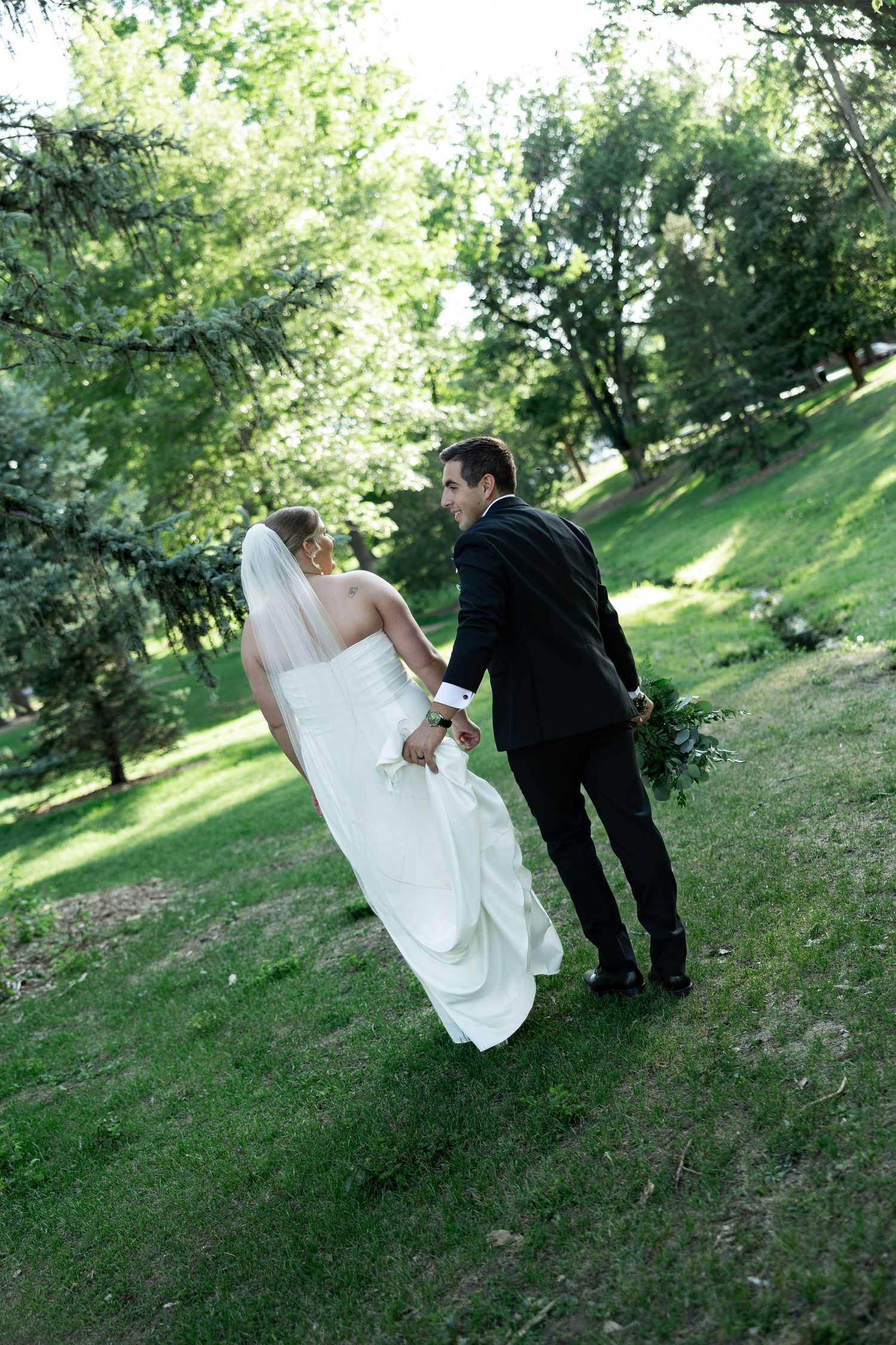 Bride and groom walking hand in hand through a lush green park, bride in white gown with veil.