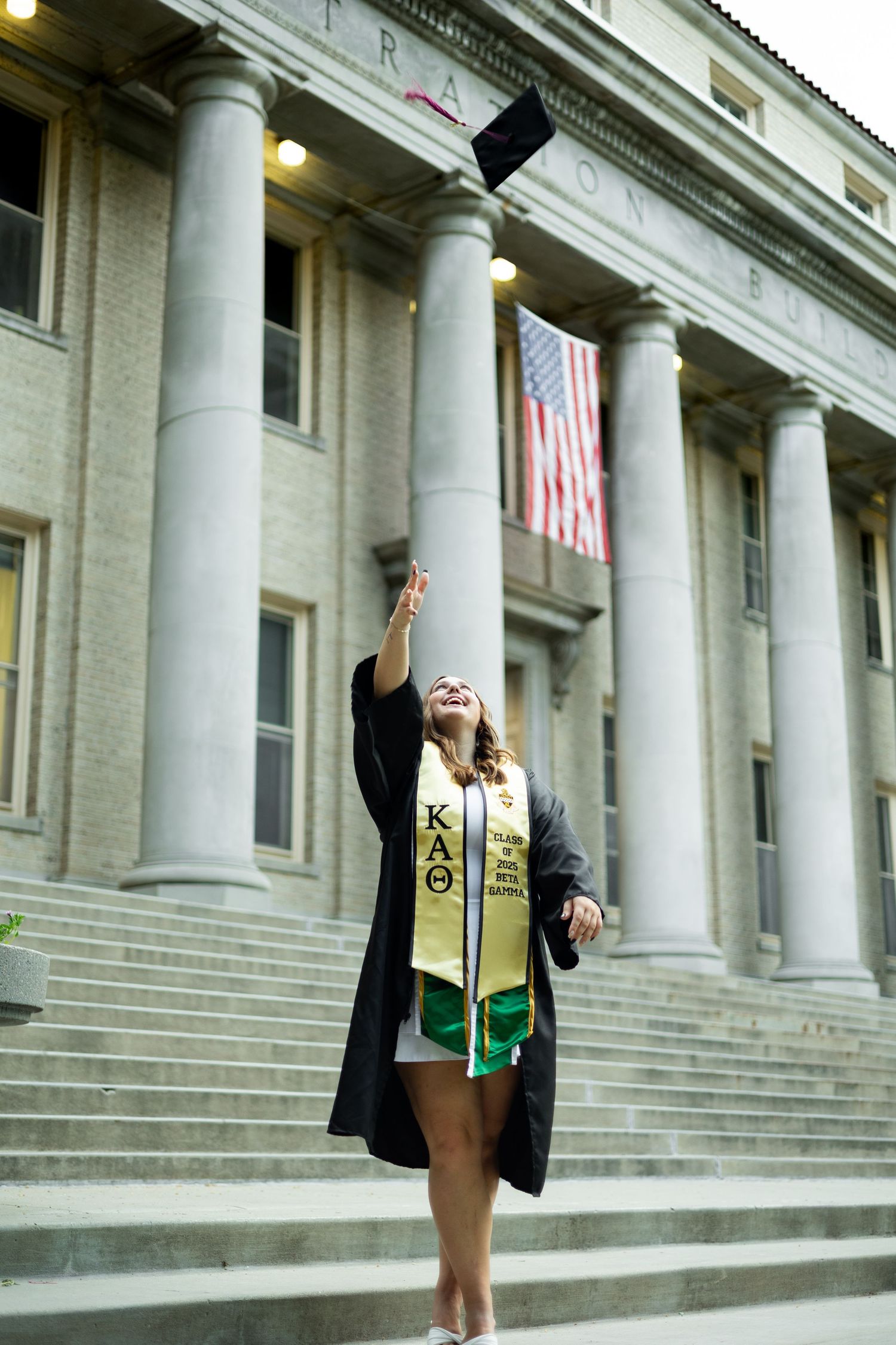 Graduate wearing cap and gown with KAO stole celebrates on steps of neoclassical building with American flag.