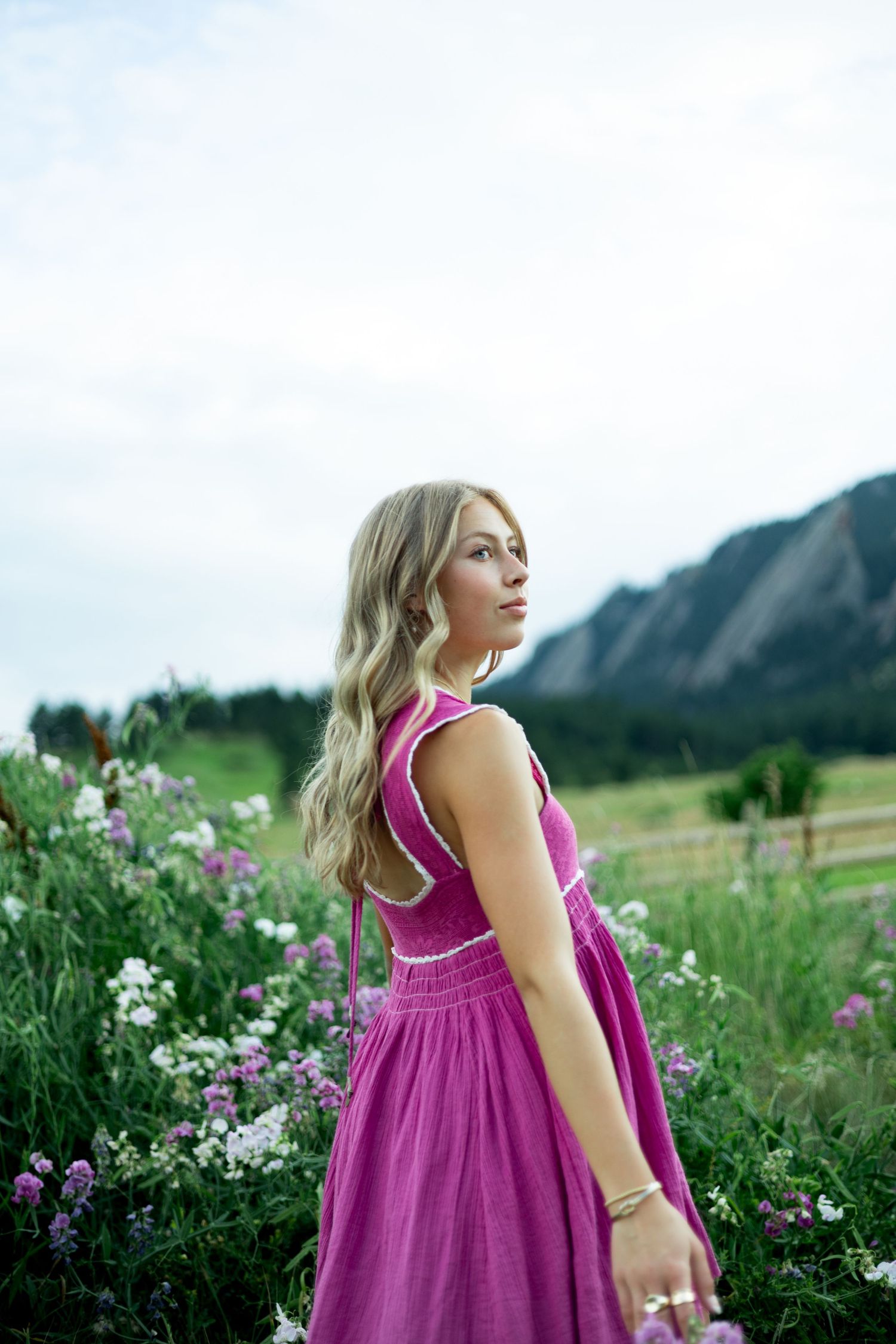 Woman in bright magenta dress stands in wildflower field with mountains and blue sky in the background.