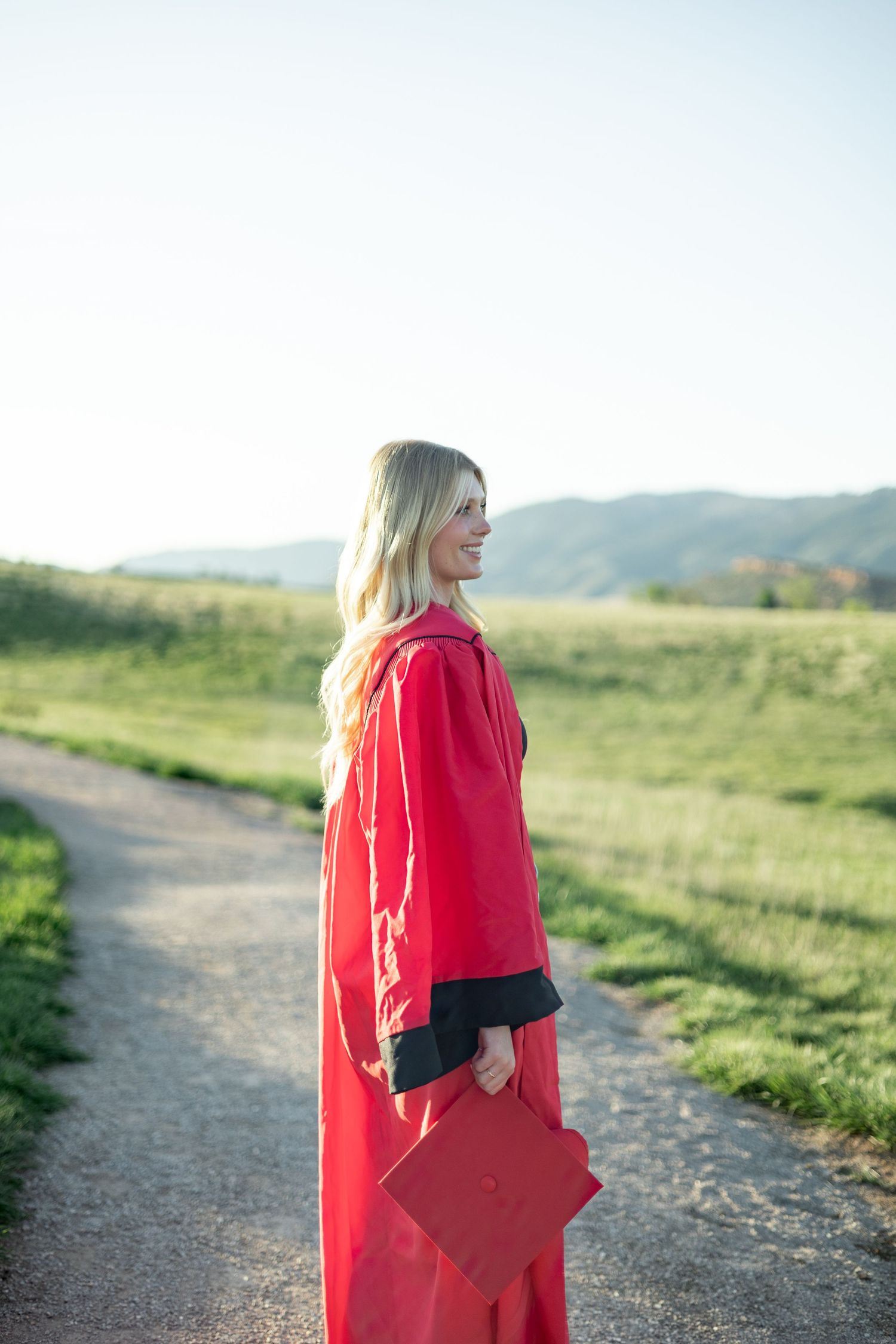 Graduate wearing a bright red graduation gown stands on a path with rolling hills in the background during sunset.