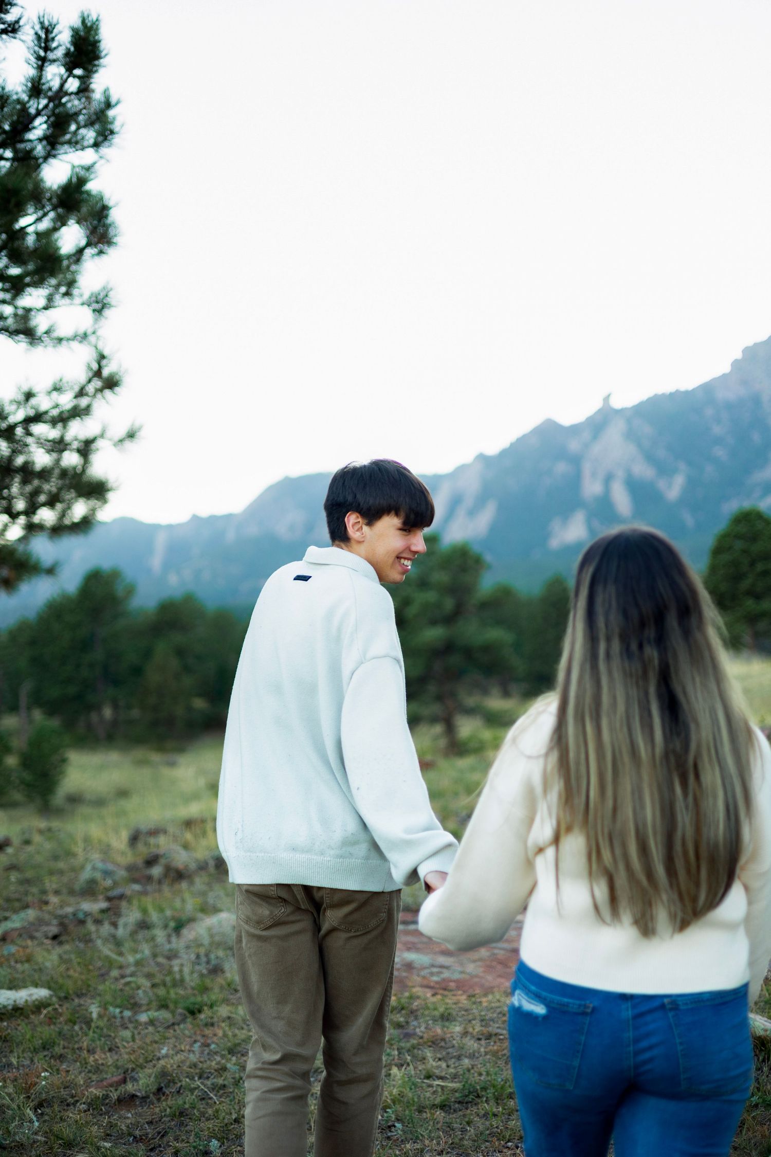 Couple holding hands in a mountain meadow, man in white sweater smiling back at woman with long hair.