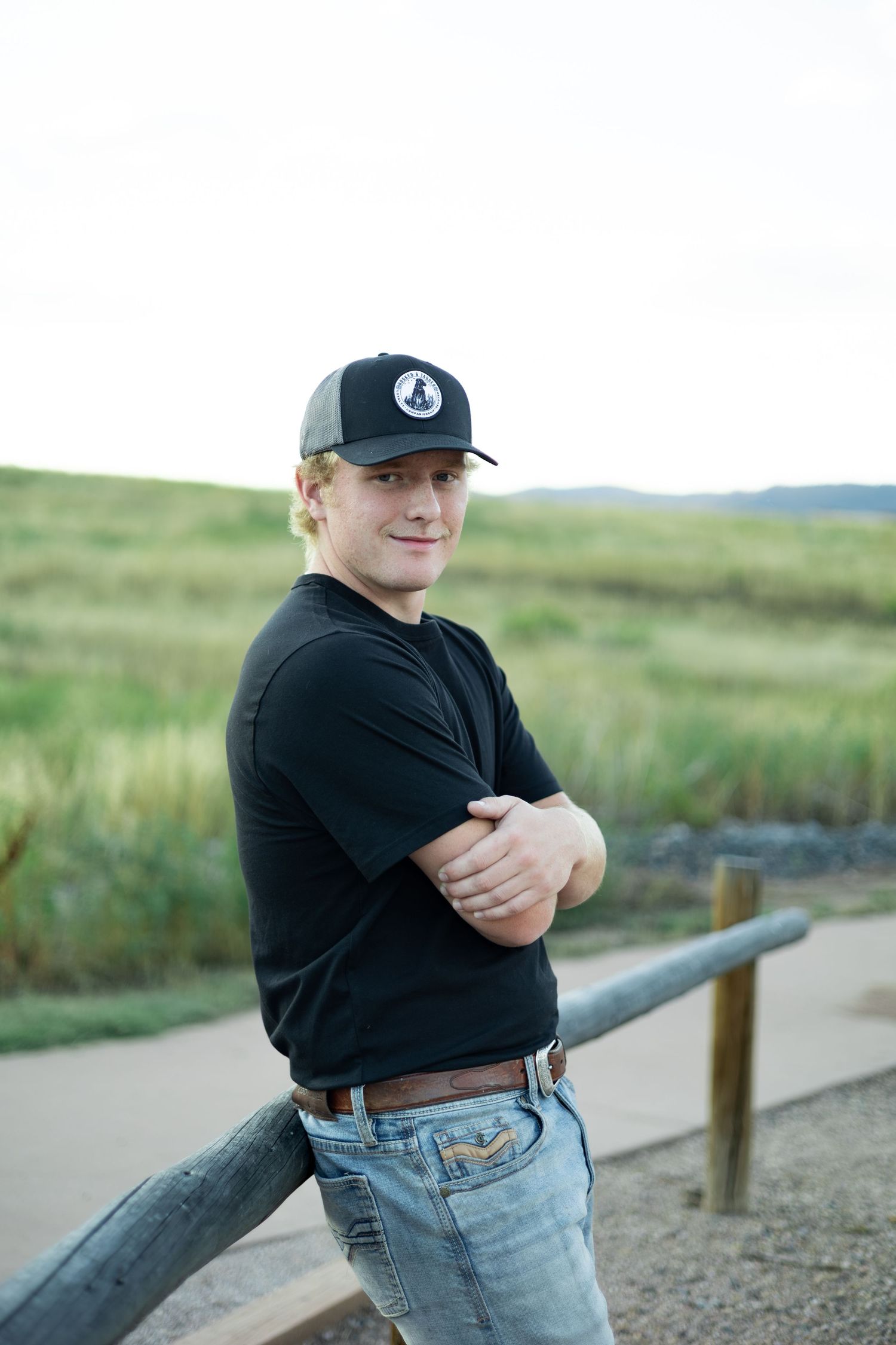 Young man in black t-shirt and cap leans on wooden fence rail outdoors with green hills in background.