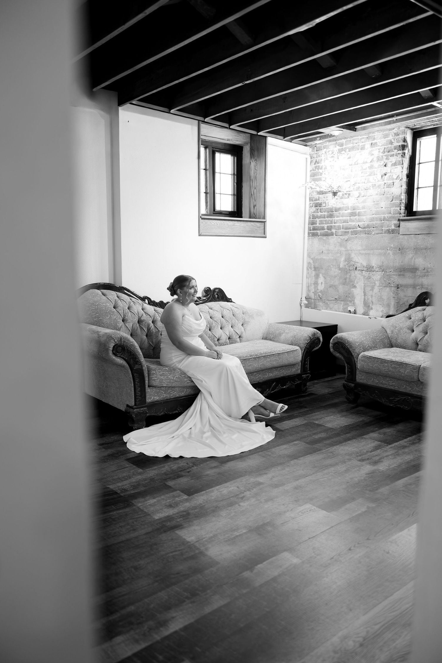 Bride in white gown sits gracefully on vintage tufted sofa in rustic brick room with hardwood floors, black and white photo.