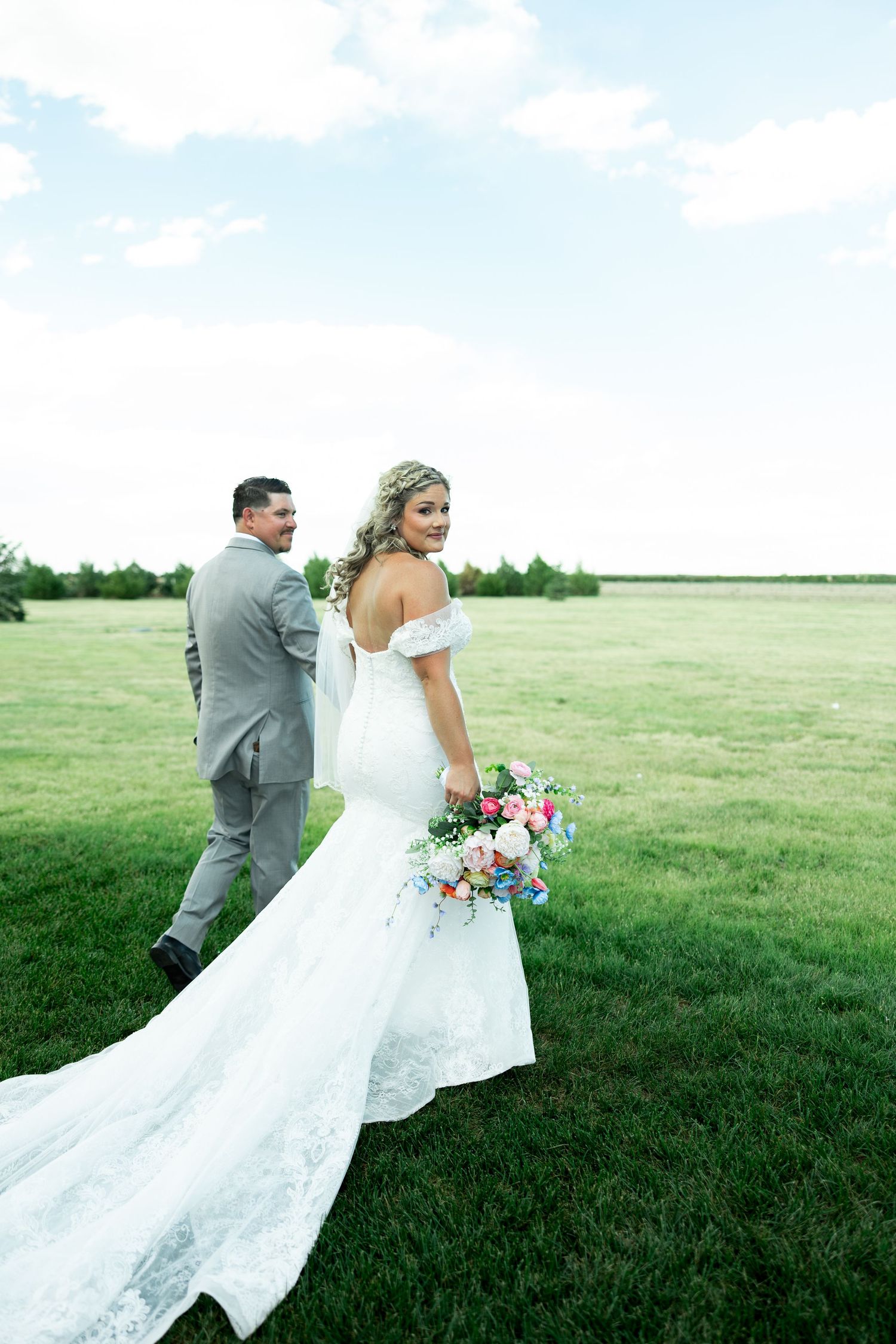 Bride in off-shoulder mermaid gown with floral bouquet walks with groom in gray suit across lush green field.