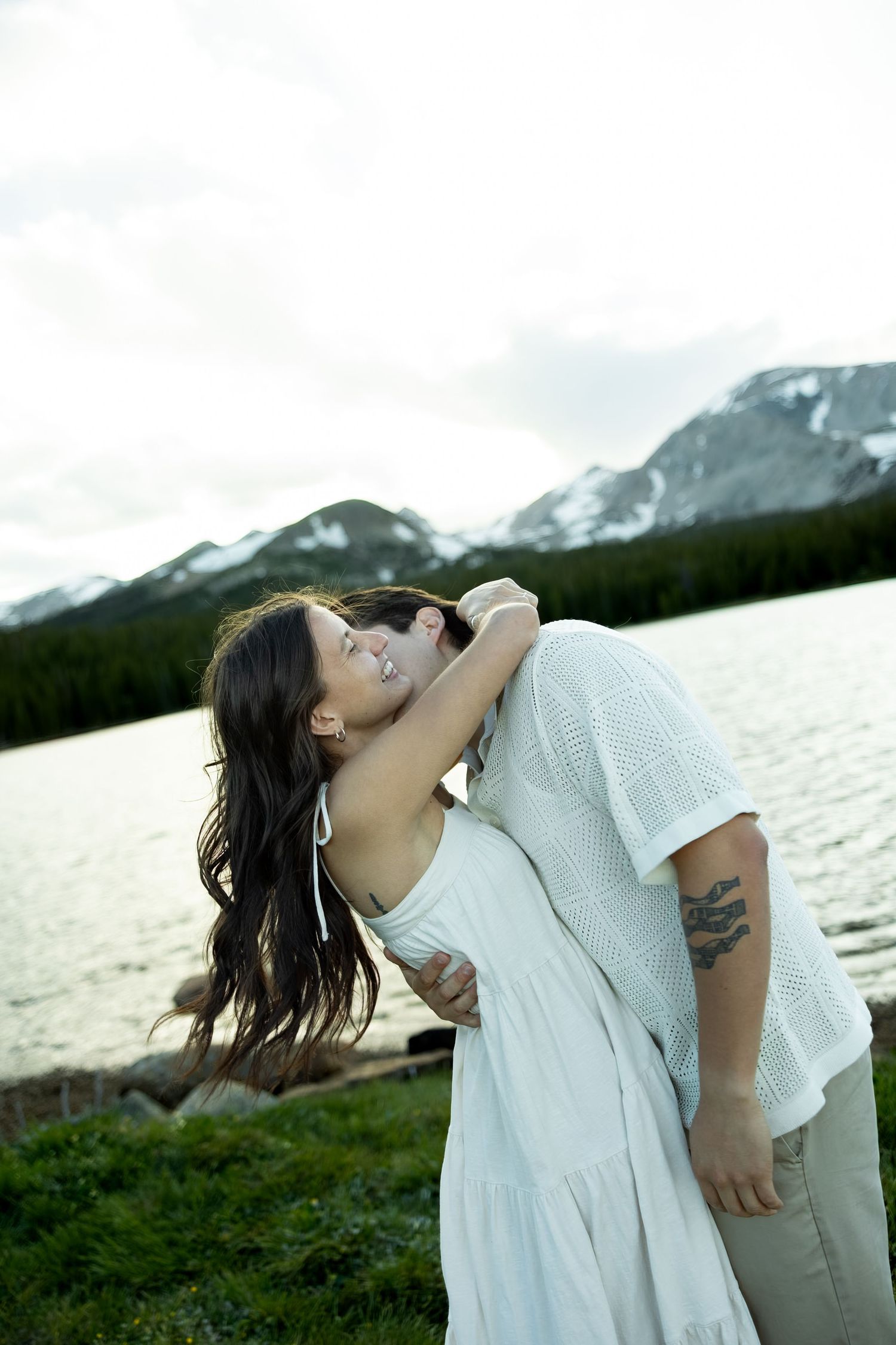 A couple shares a romantic kiss by a mountain lake, the woman in a white dress with long dark hair, the man with a tattoo.