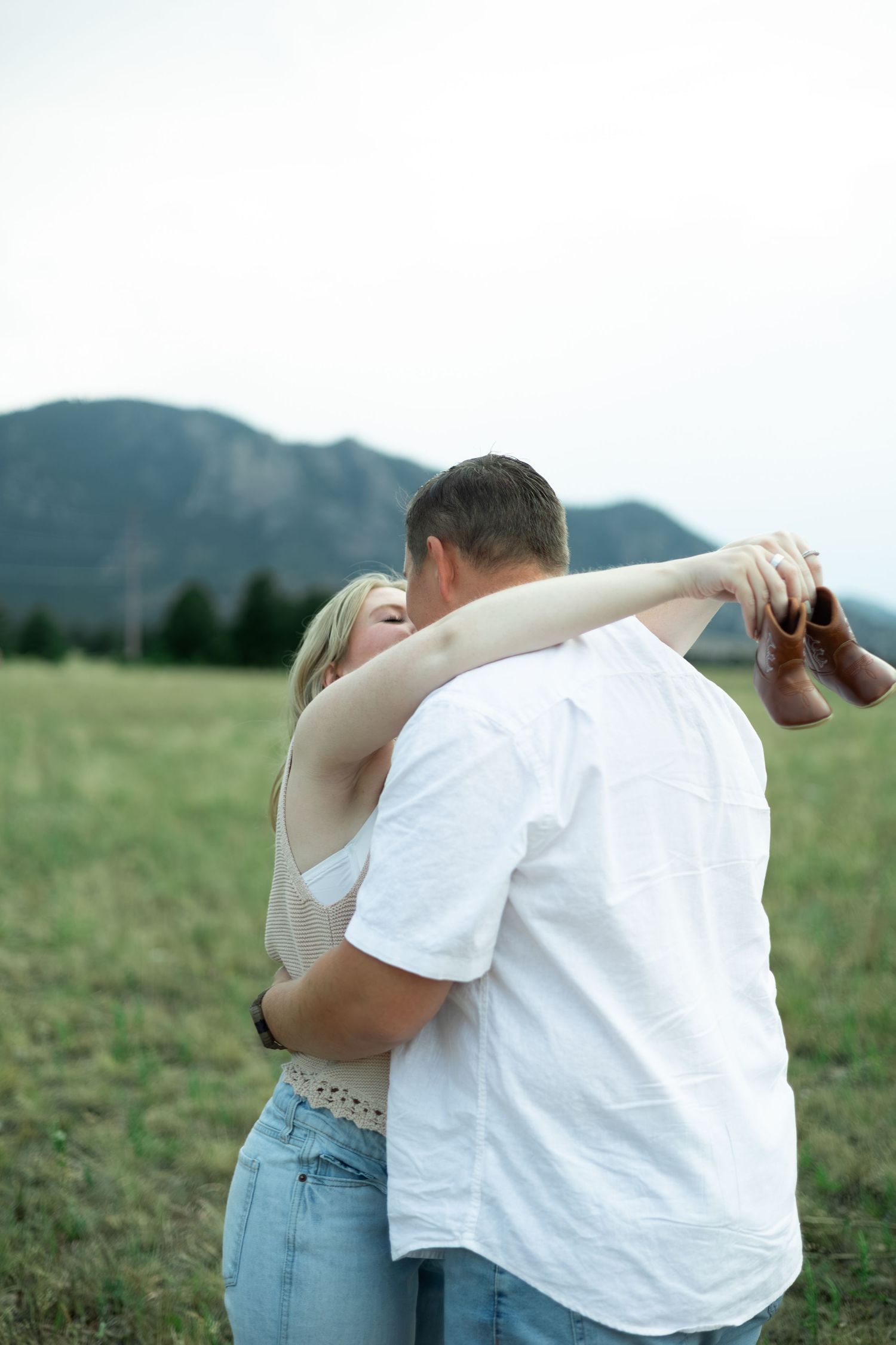 Two people in casual attire share a tender embrace while standing in a scenic grassy meadow with mountains in the background.