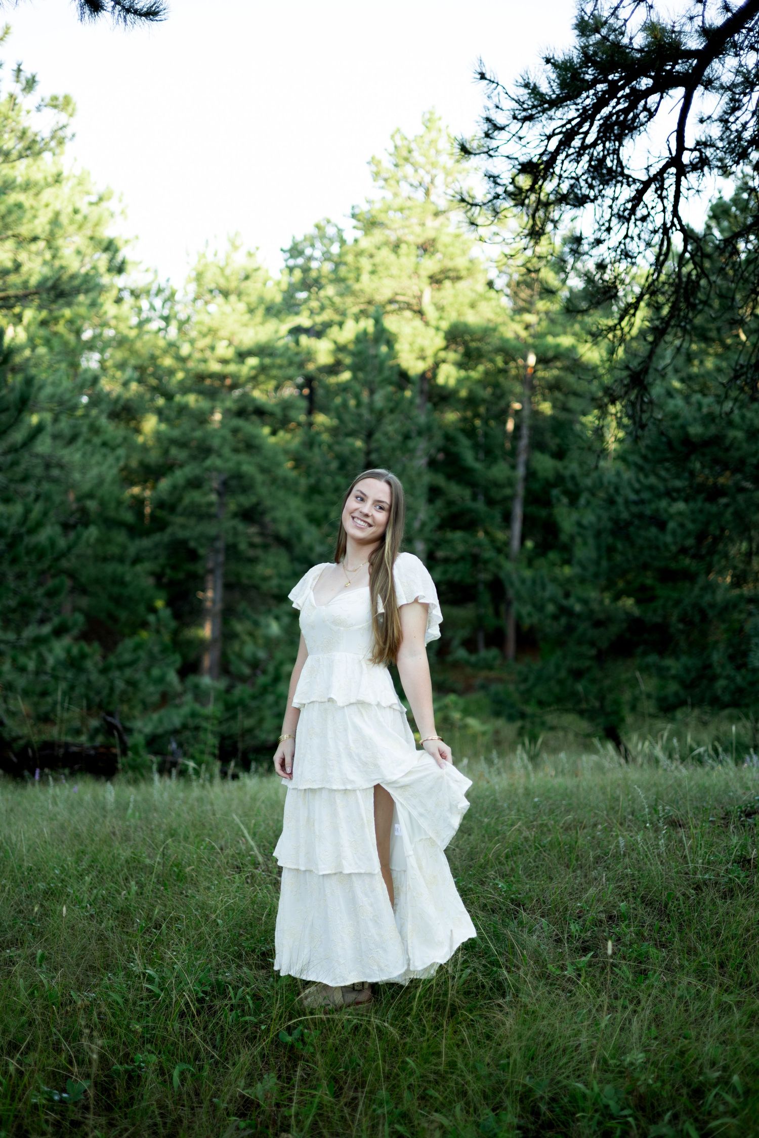 A person in a flowing white dress twirls playfully in a sunlit forest meadow surrounded by tall pine trees.