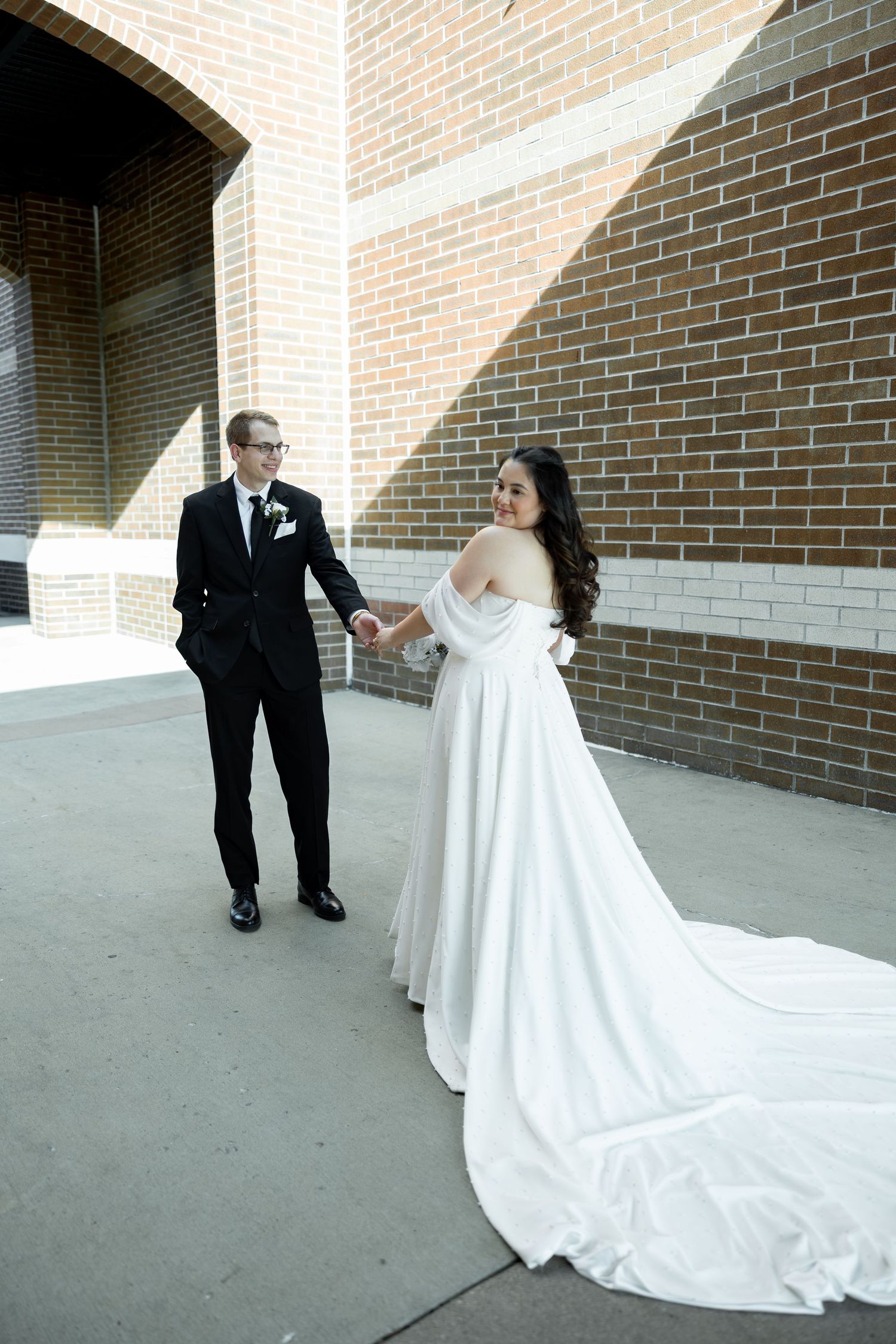 Bride in white gown and groom in black suit hold hands outside brick building.
