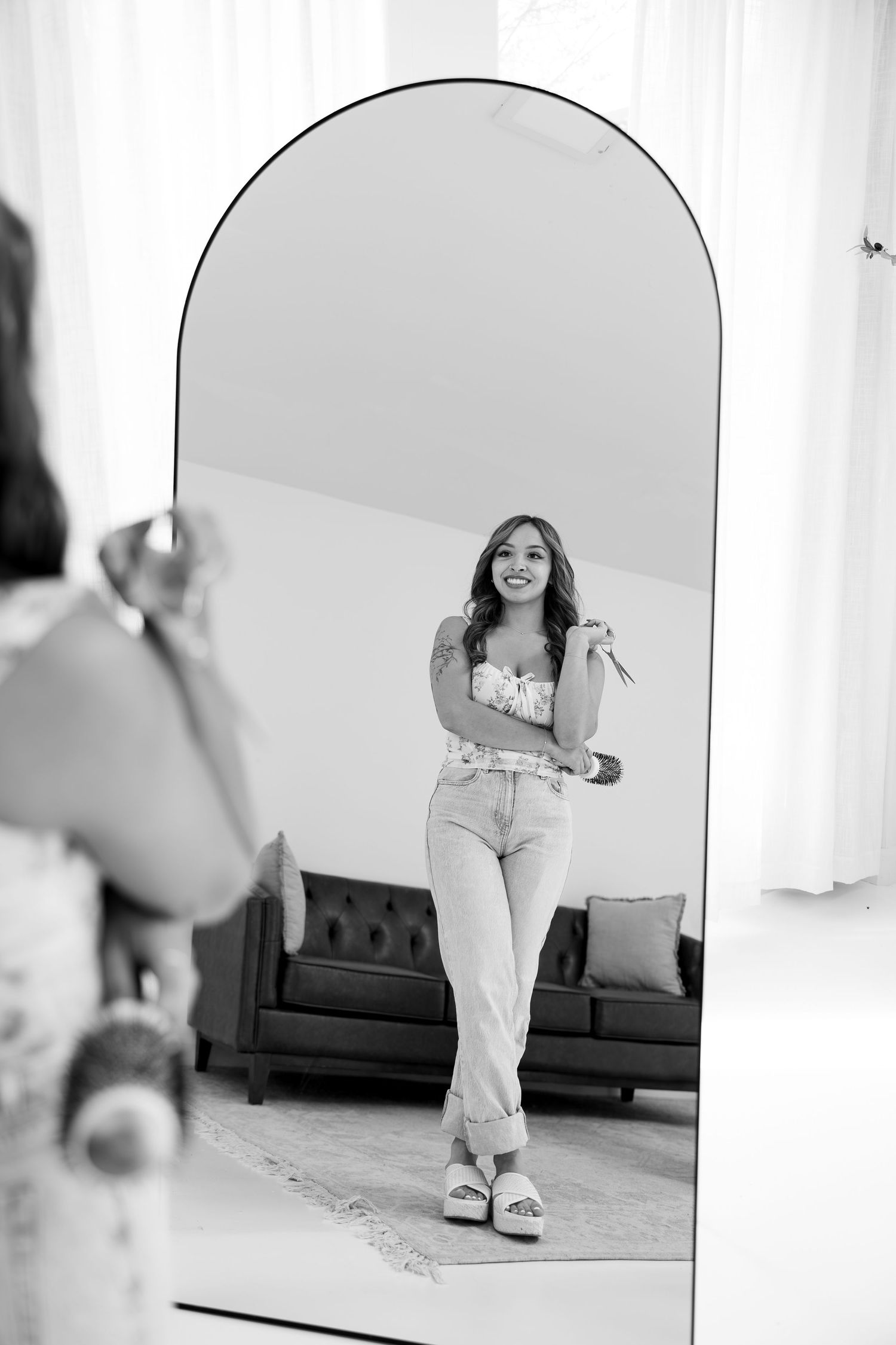 Black and white photo of a smiling woman in jeans posing in front of a large arched mirror.