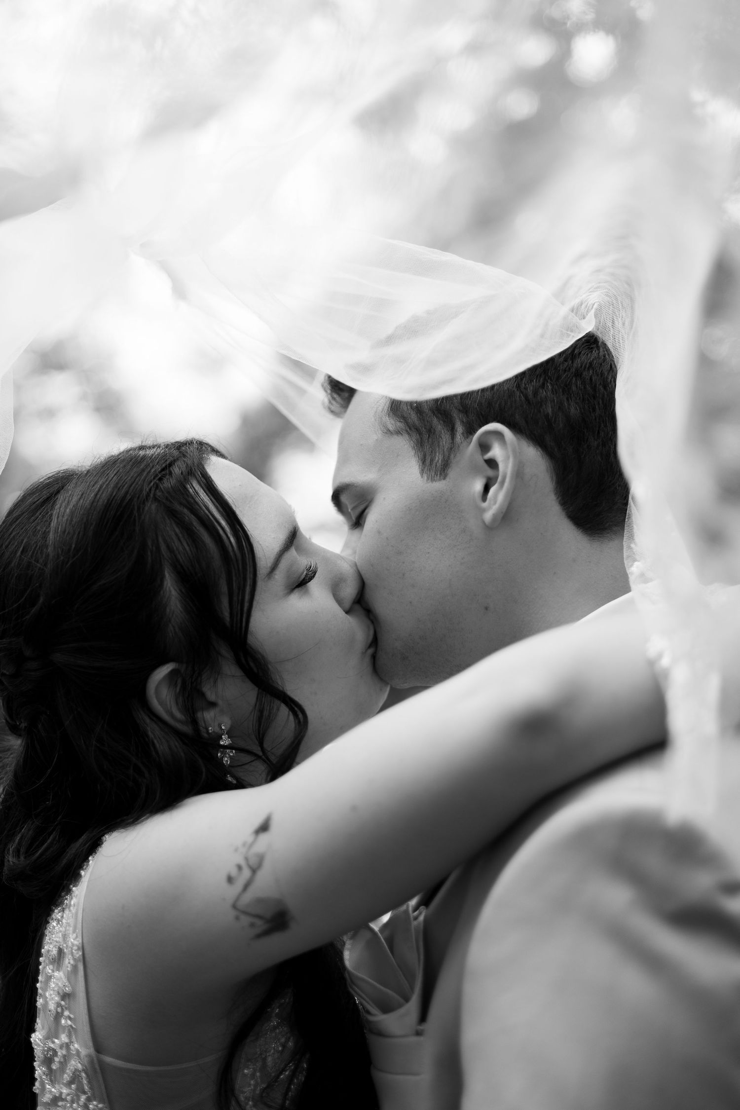 A romantic black and white photo captures a newlywed couple sharing their first kiss under a flowing veil.