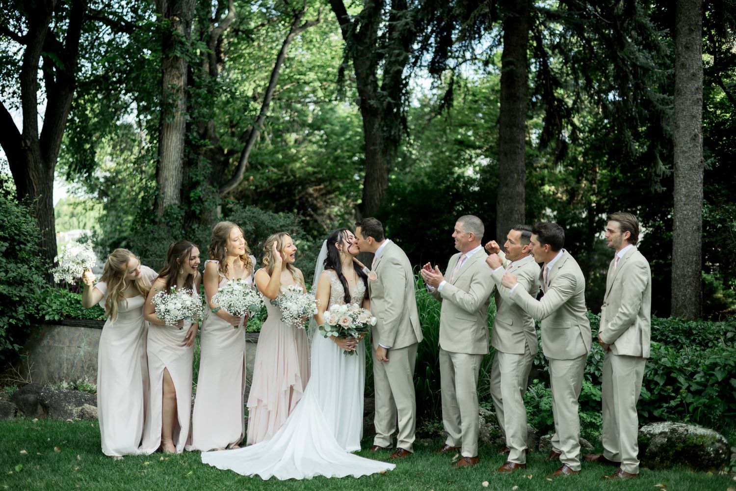 A wedding party dressed in beige and white stands beneath towering trees in a lush forest setting.