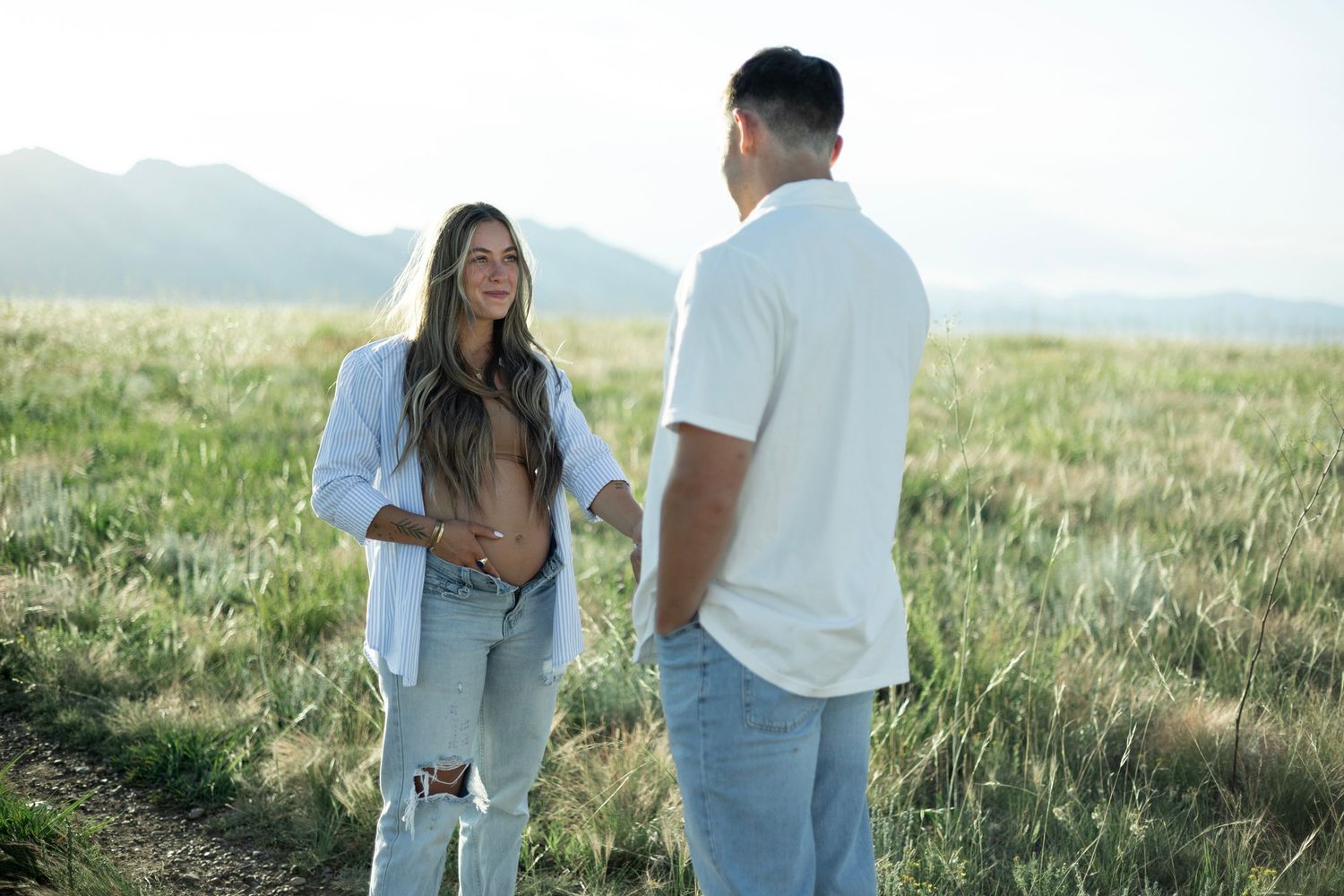 Two people in casual white outfits having a conversation in a grassy field with mountains in the background.
