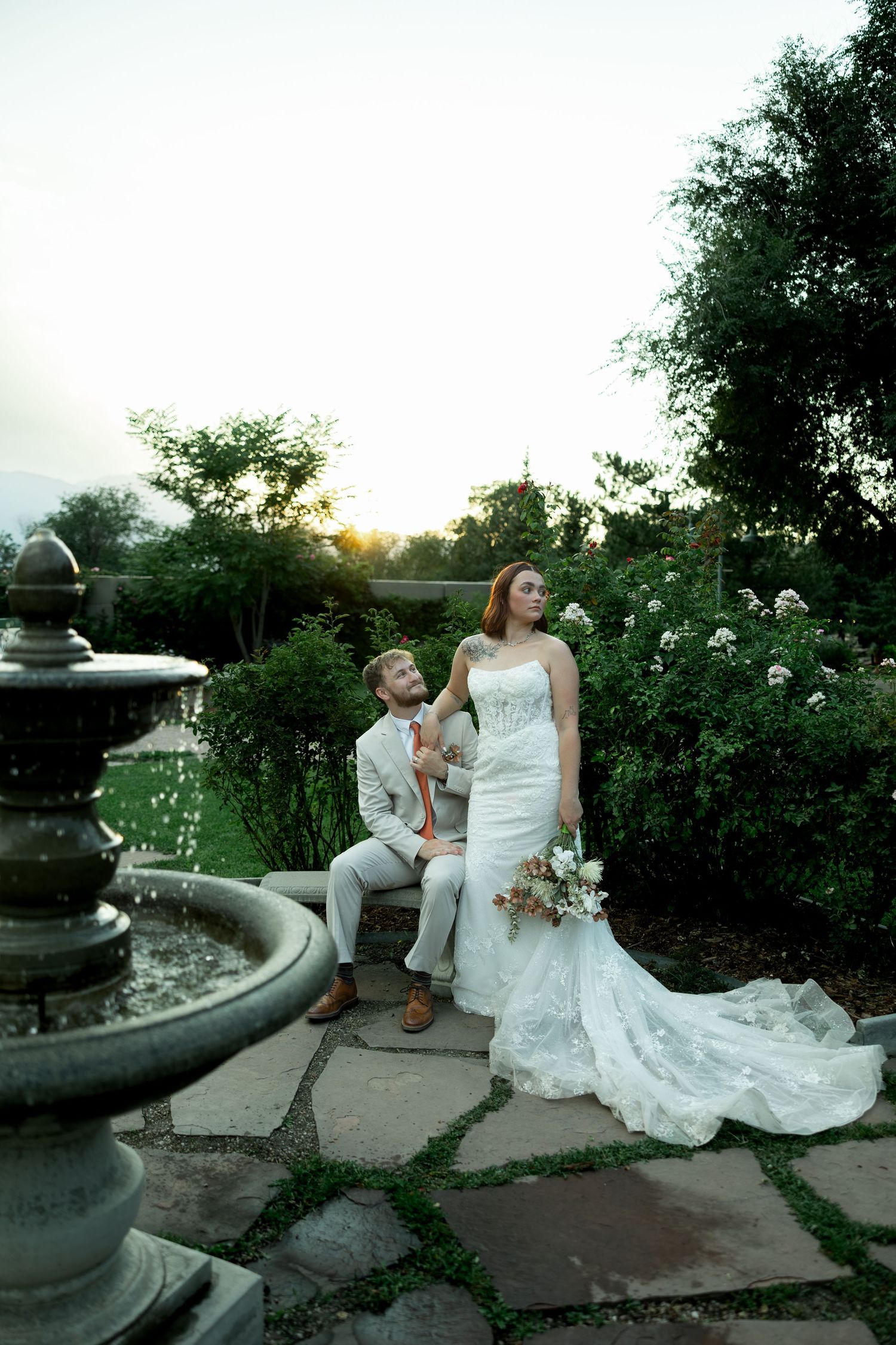 Bride and groom share romantic sunset moment by garden fountain, bride in lace gown with long train.