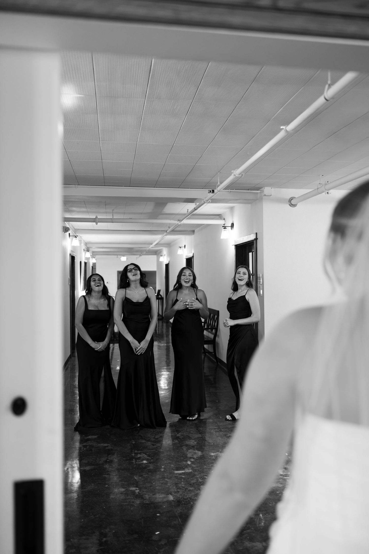 Bridesmaids in black gowns react with awe seeing bride in hallway, black and white wedding photo.