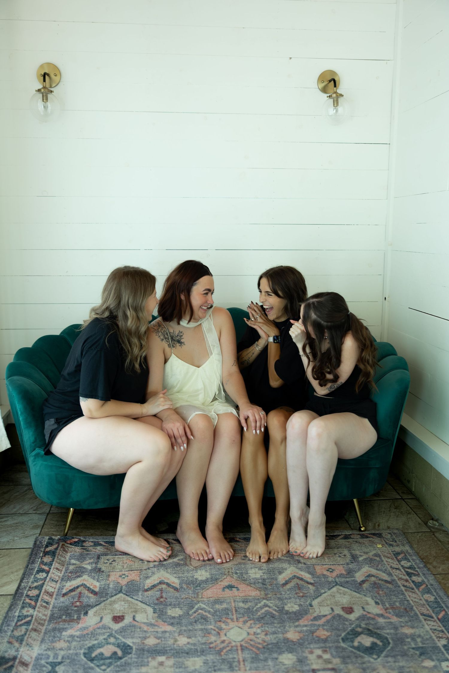Four women sitting closely together on a teal velvet sofa, laughing and sharing secrets in a cozy room.