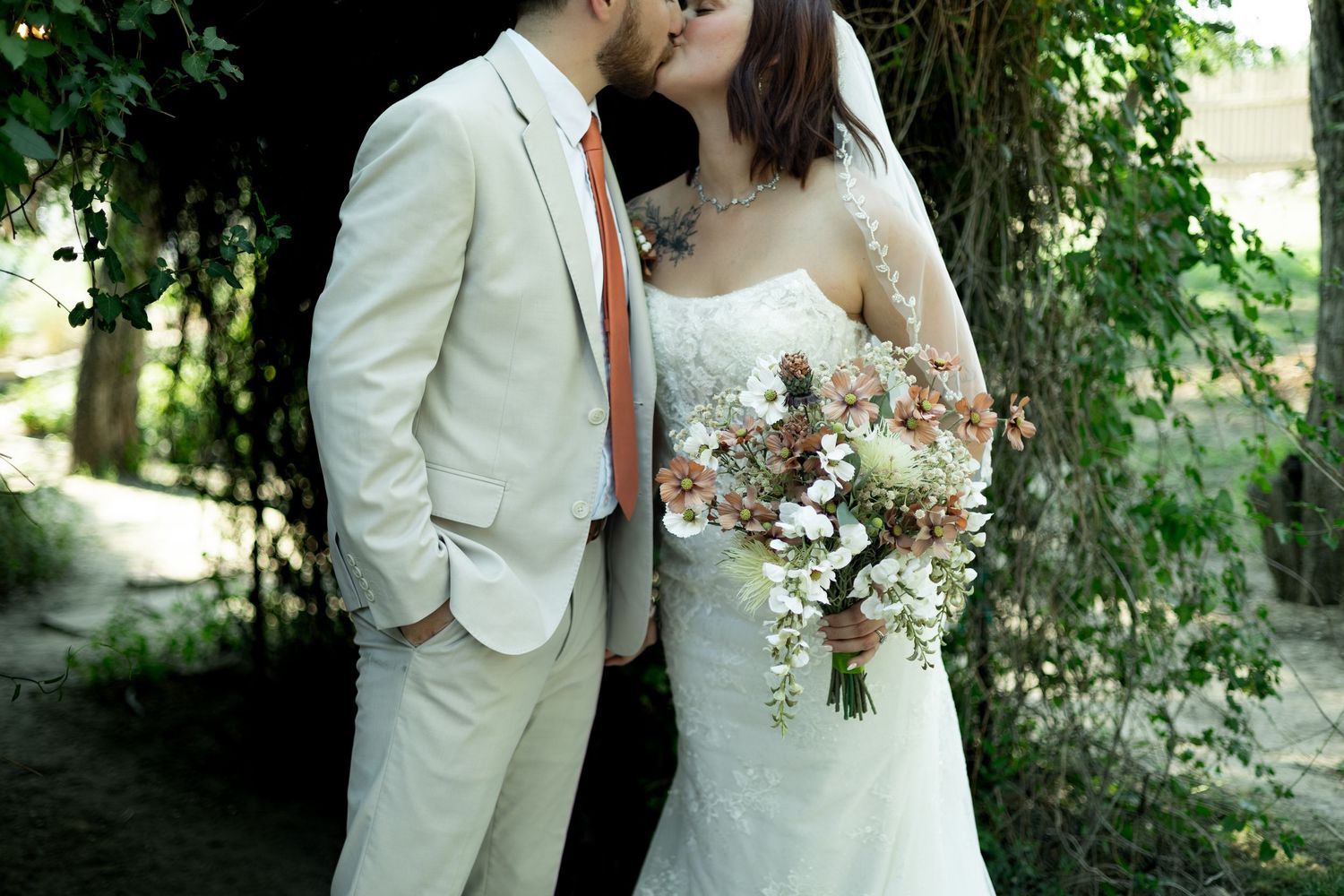 Bride and groom share a kiss outdoors, she holds a boho bouquet, he wears a beige suit with orange tie.