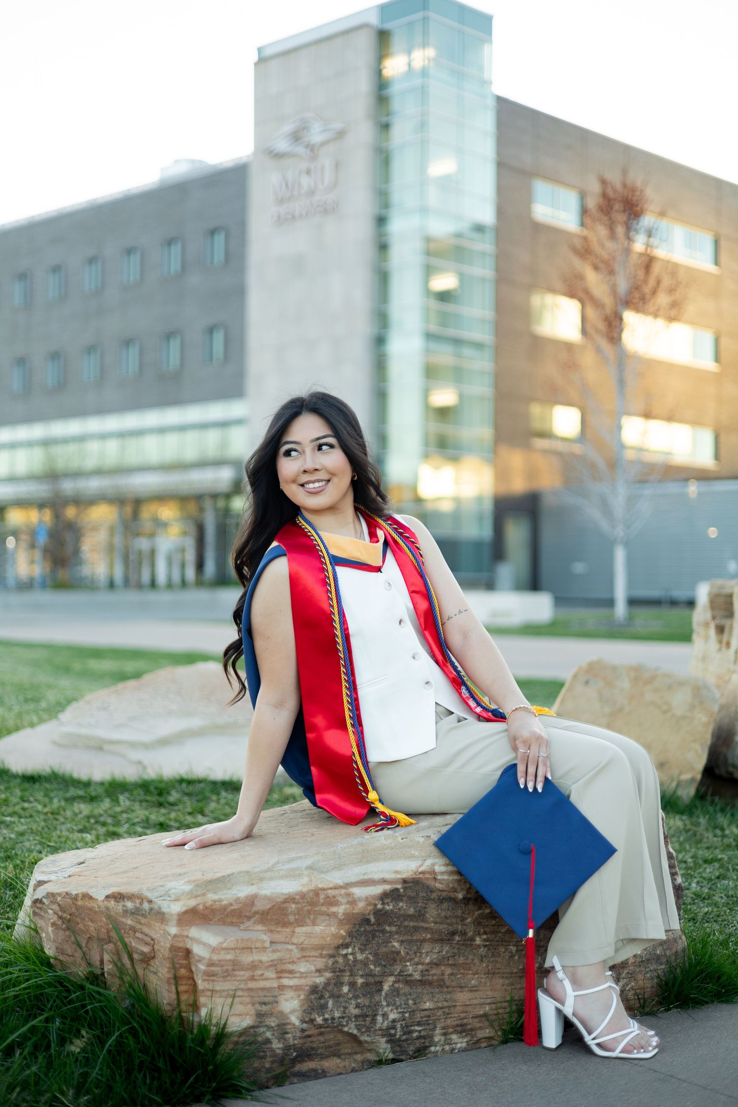 Graduate in red graduation stole sits on a rock outside a modern university building during commencement season.