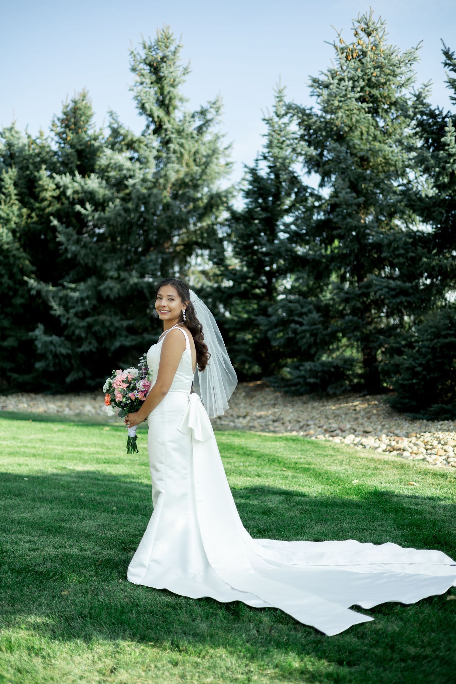 A serene wedding portrait series showing a bride in a flowing white gown resting gracefully on green grass near evergreen trees.