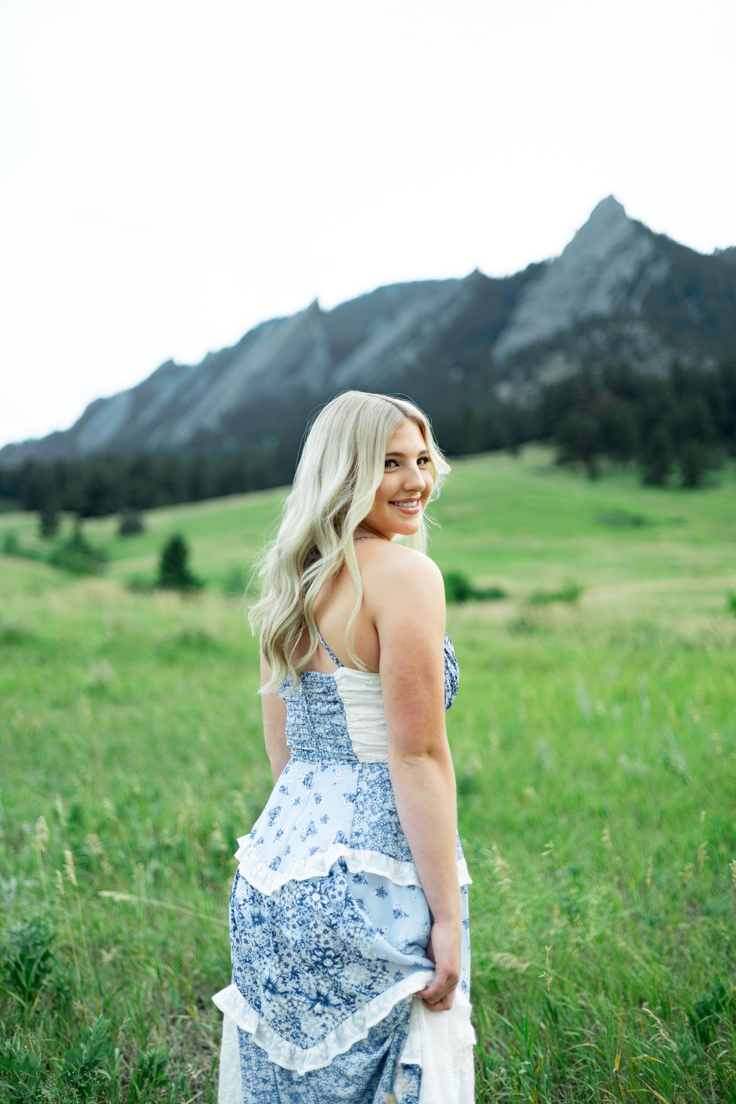 Woman in flowing blue floral dress poses against scenic mountain backdrop in green meadow.