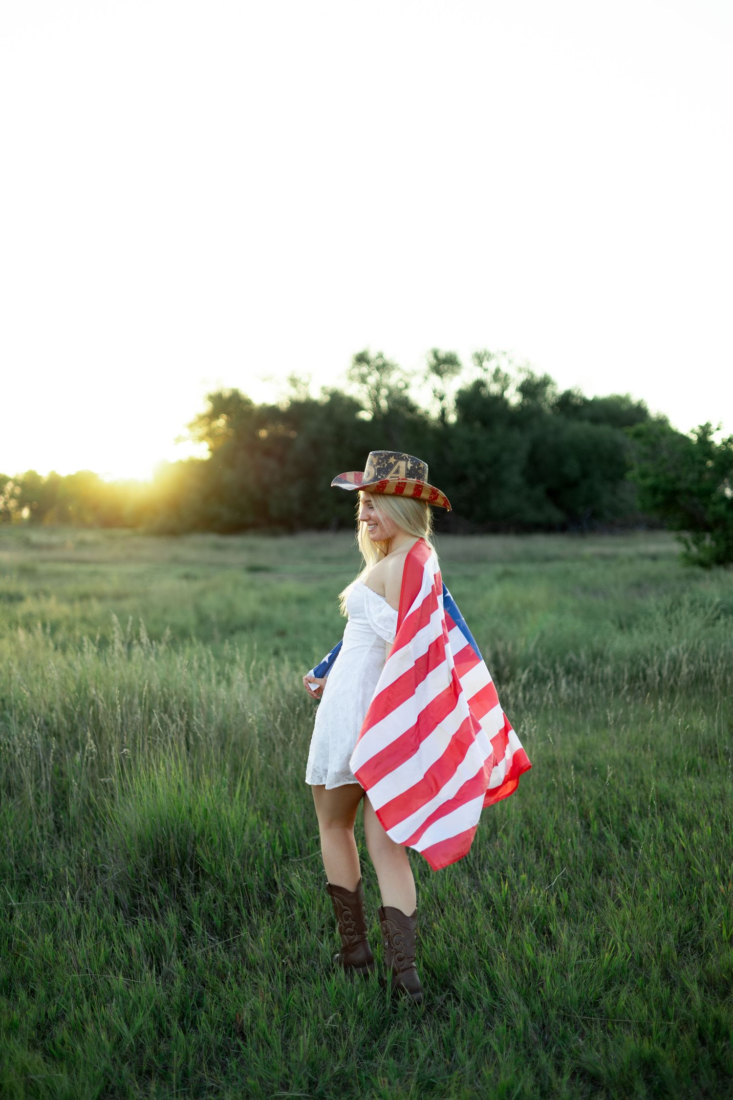 Blonde woman in cowboy hat draped in American flag standing in green field at golden sunset.