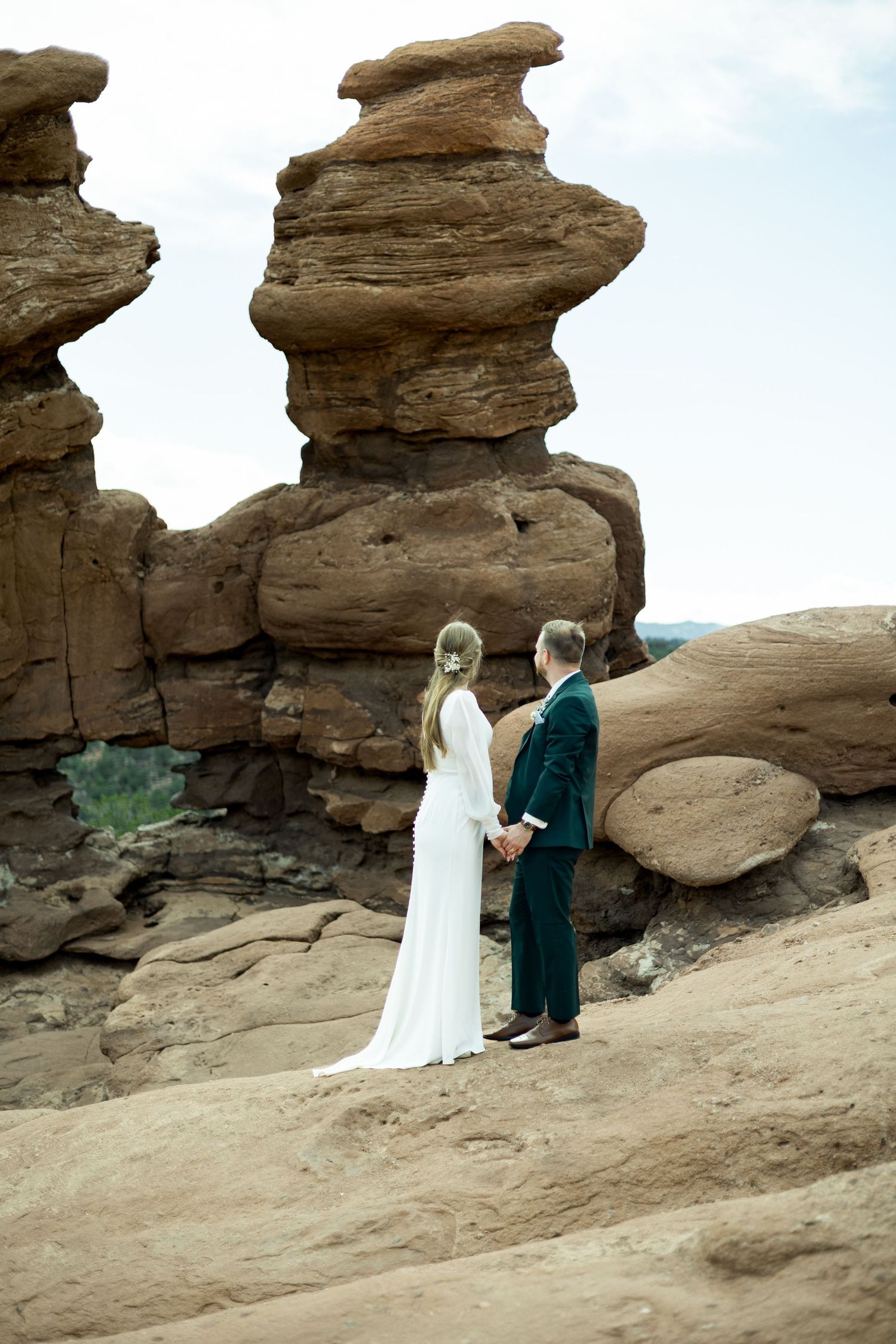 Two figures in formal attire stand together against dramatic red rock formations in a desert landscape.
