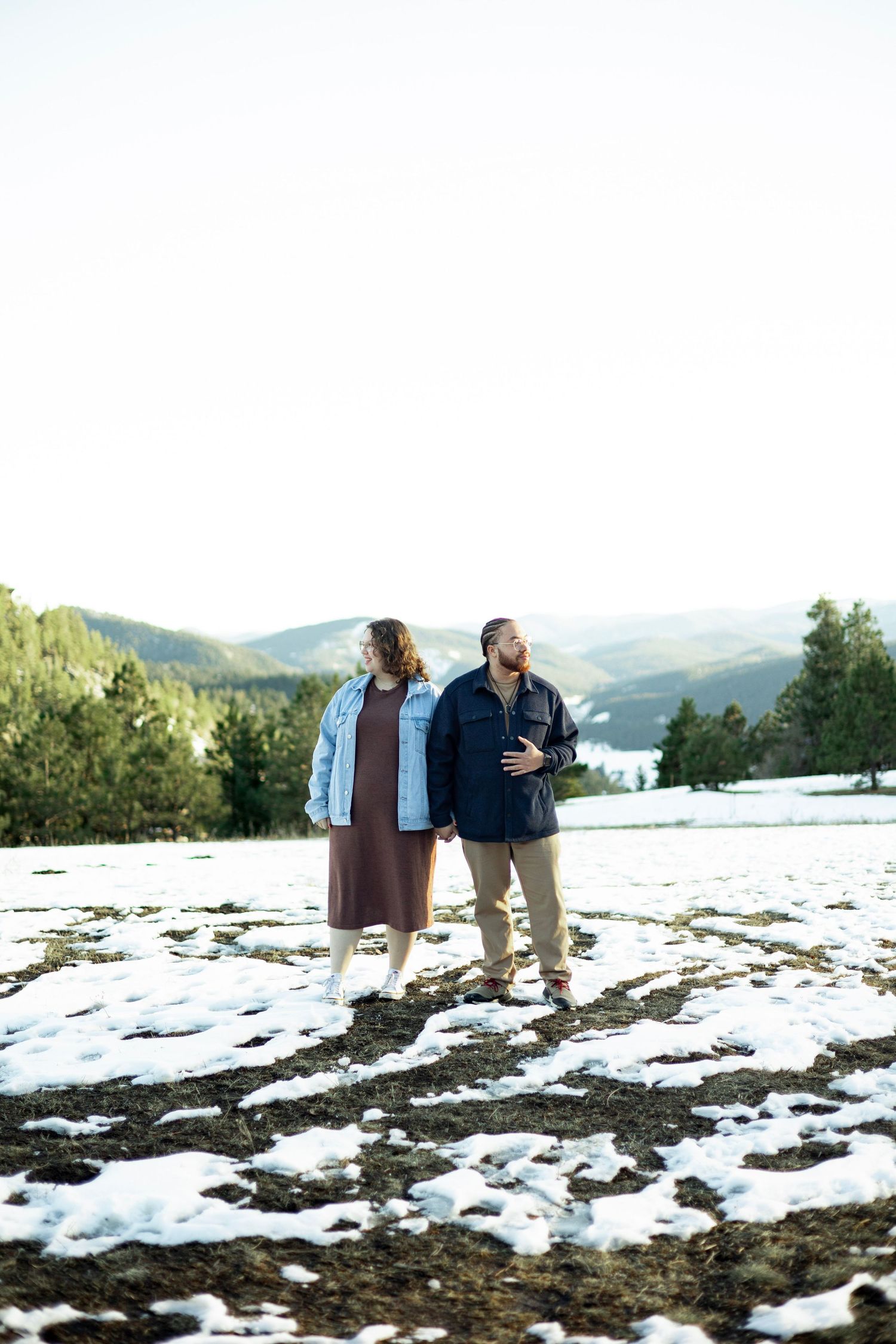 Couple standing together on a snowy mountain landscape with pine trees and hills in the background on a sunny day.