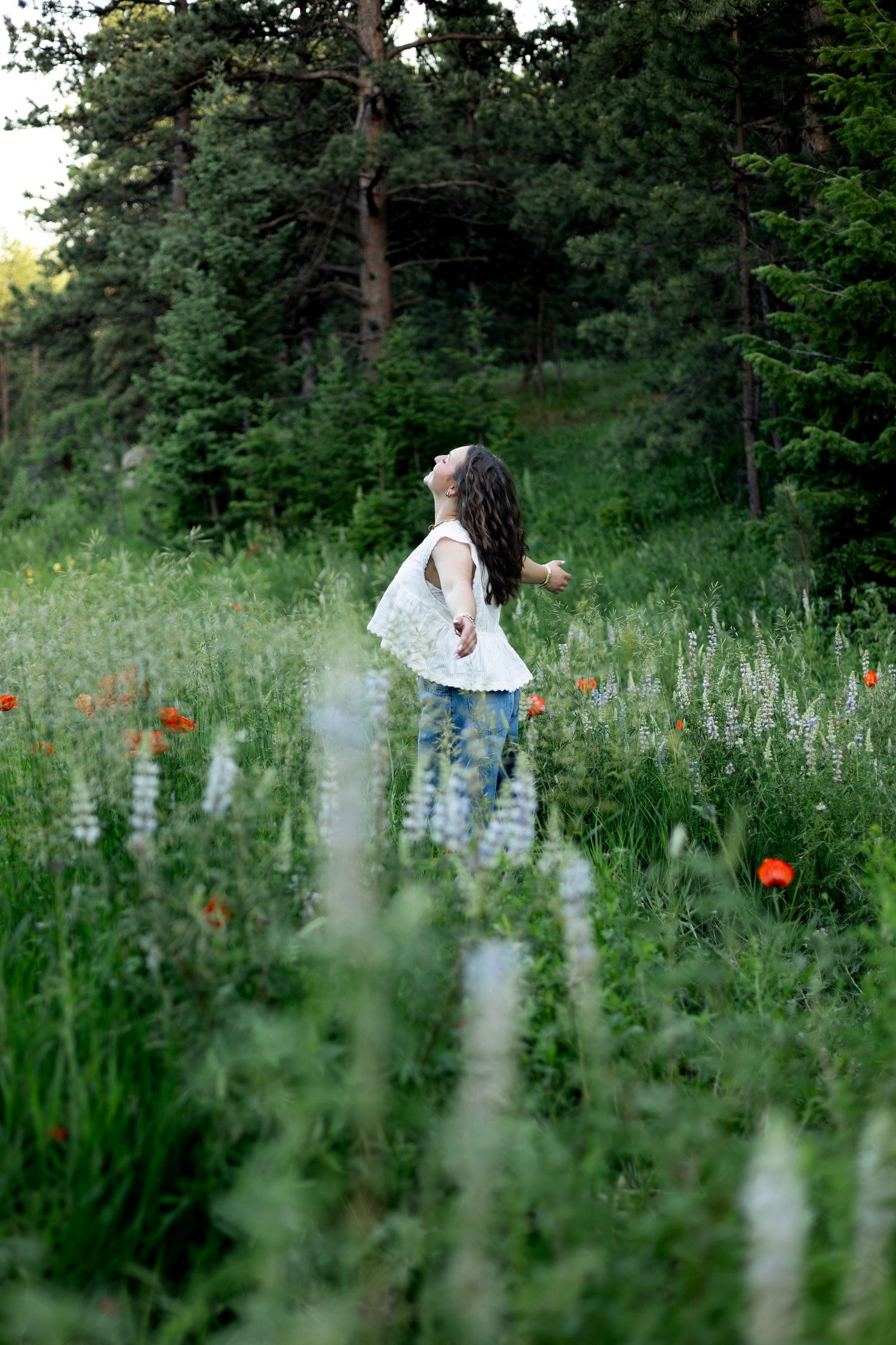 Young girl in white top and jeans twirling joyfully in a wildflower meadow with red poppies and green forest backdrop.