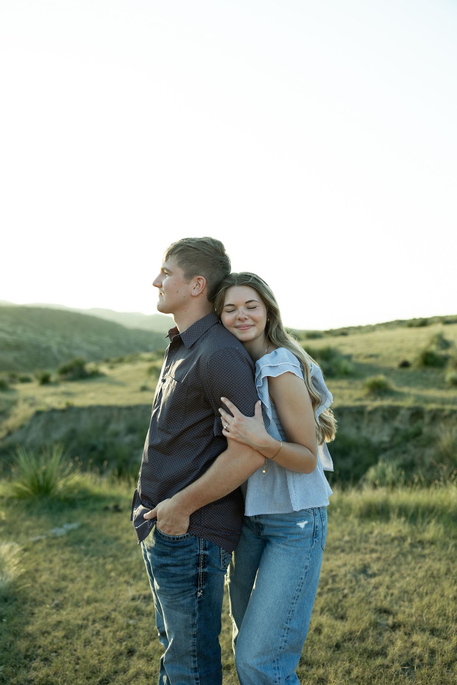 A couple embraces outdoors in a green field, woman hugging man from behind, both wearing jeans, smiling peacefully.