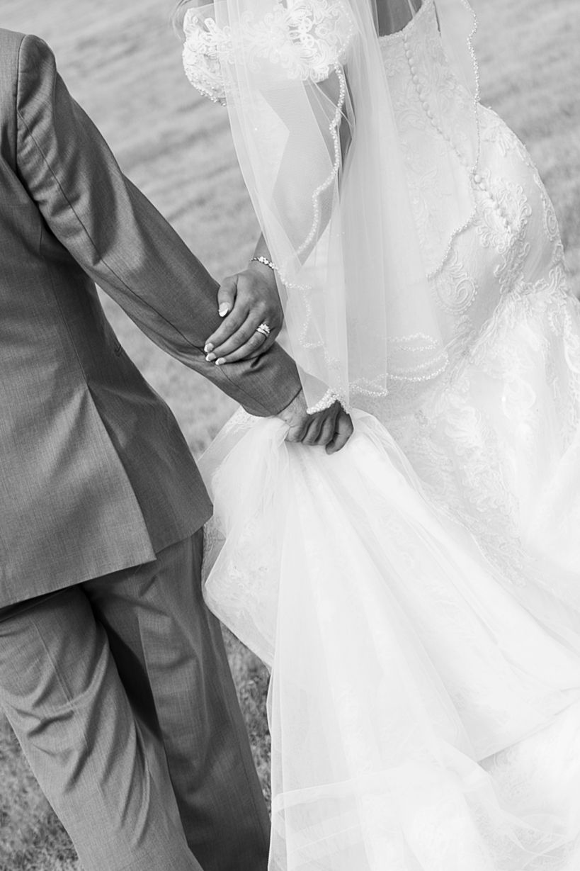 Black and white photo of bride and groom holding hands, showcasing wedding rings and elegant bridal gown.