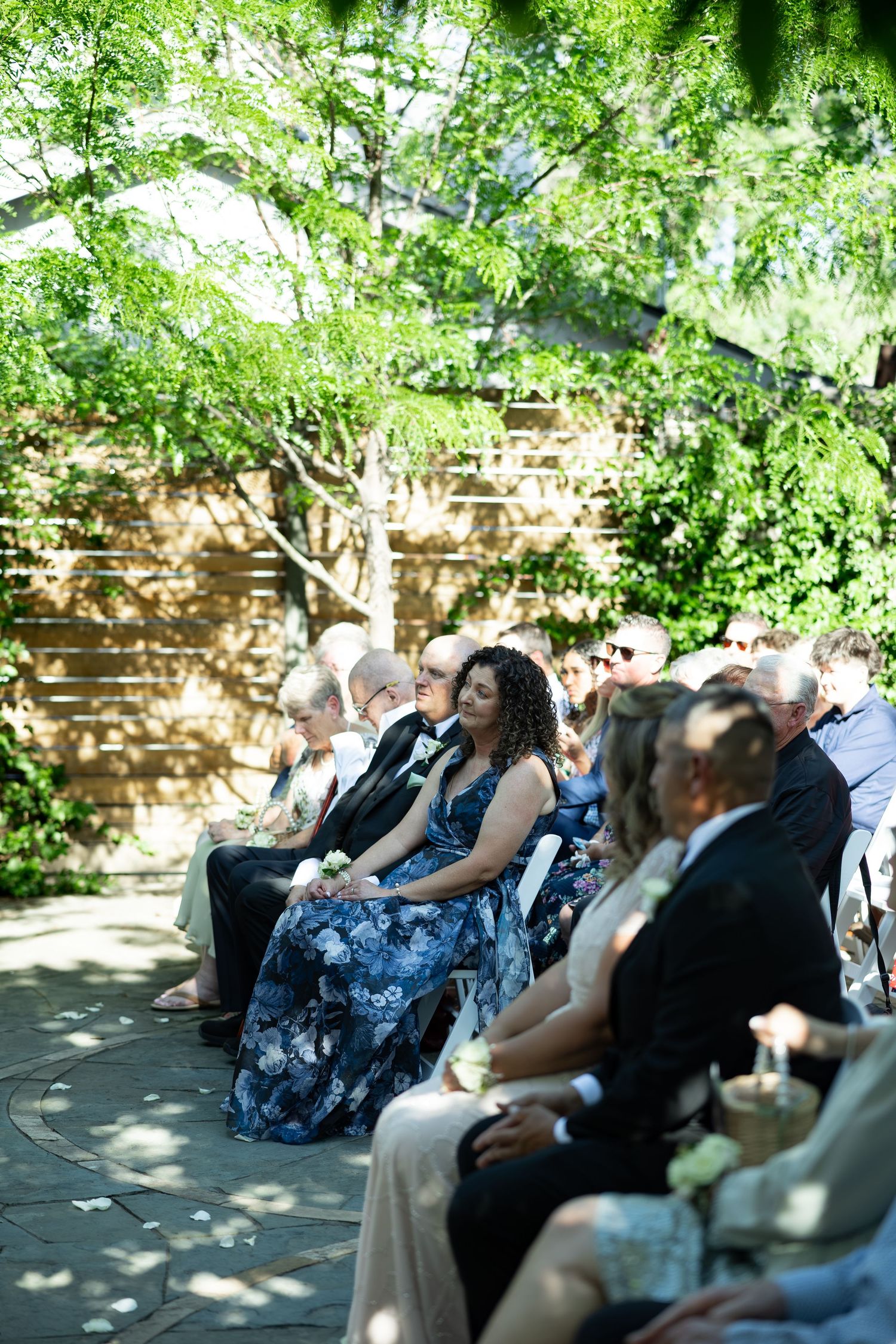Wedding guests seated outdoors in white chairs, woman in floral blue dress in focus, surrounded by green trees.