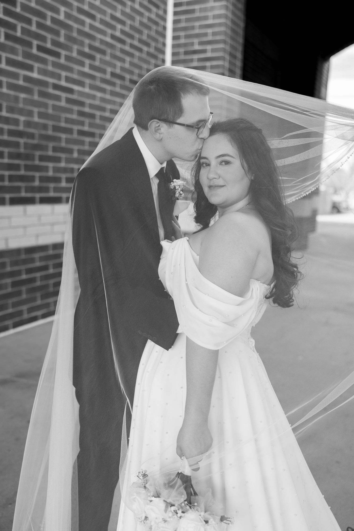 Black and white wedding photo of groom kissing bride on forehead under veil, bride holding bouquet near brick wall.