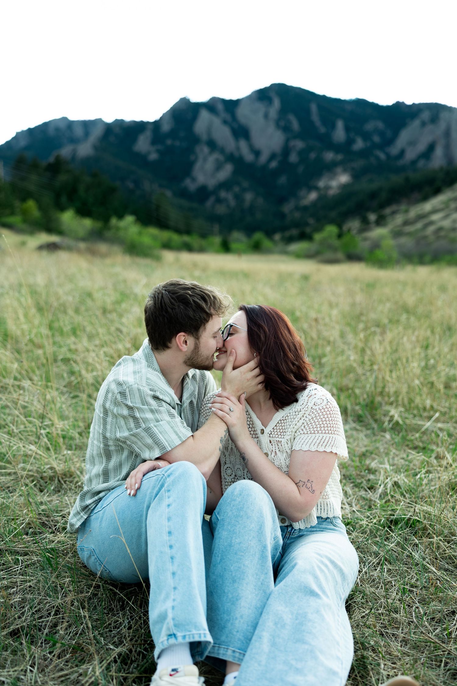 Couple sharing a romantic kiss while sitting in a grassy field with stunning rocky mountains in the background.